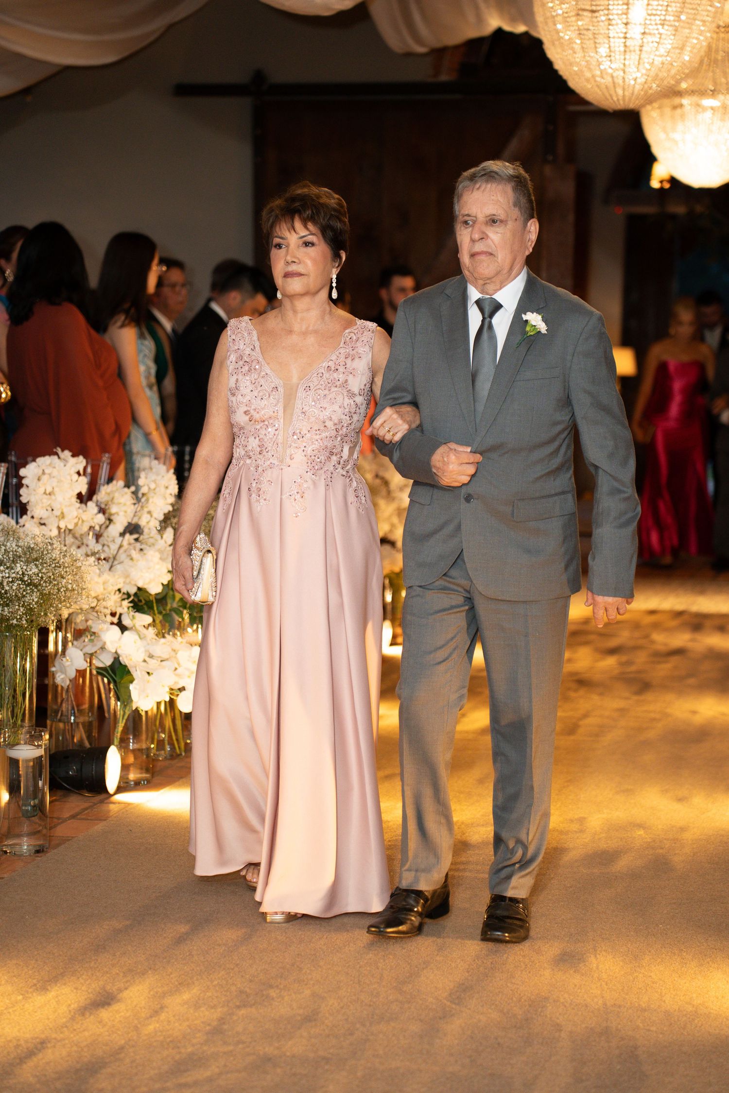 Elegant couple walking down wedding aisle, woman in pink gown, man in gray suit with boutonniere.