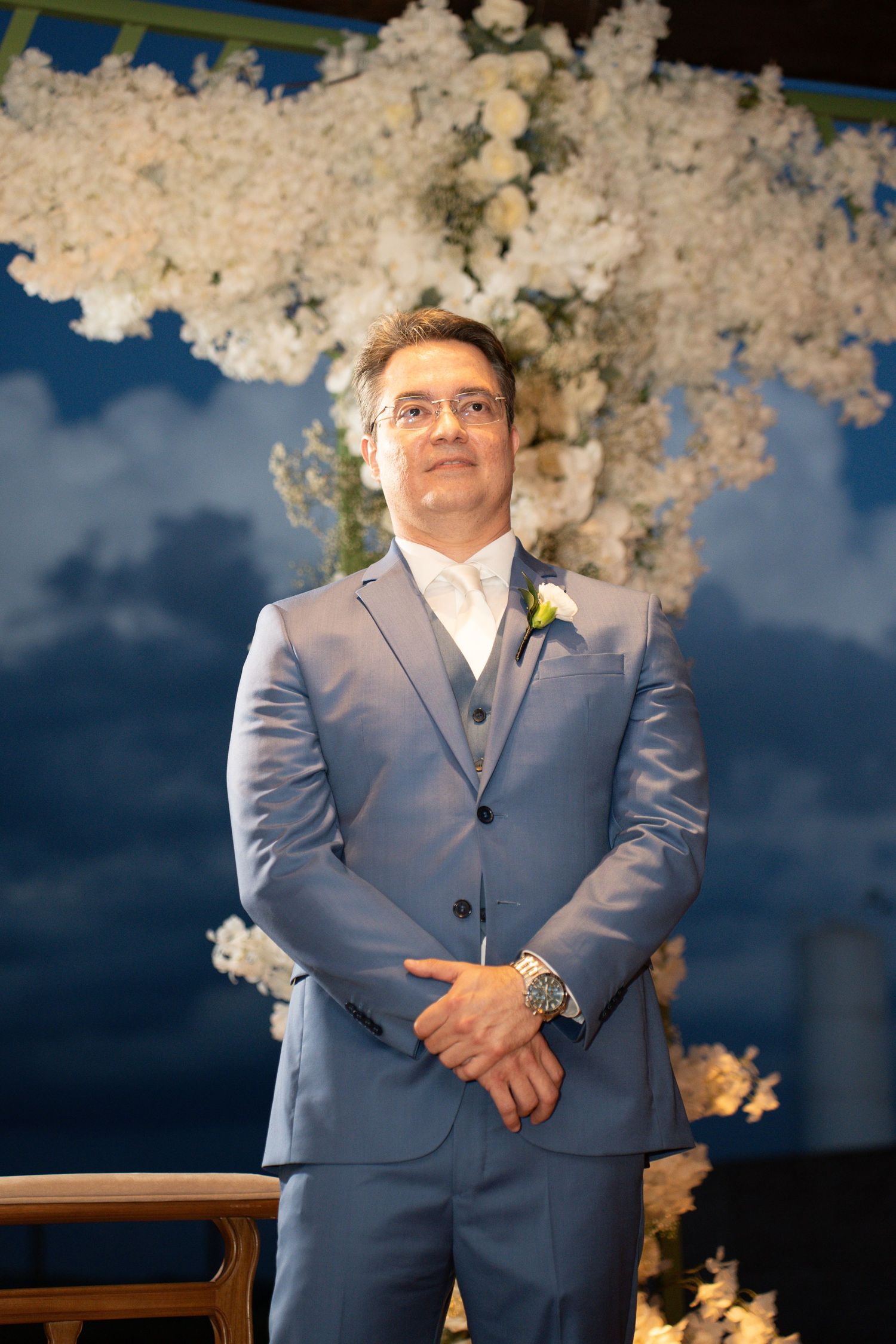 Groom in blue suit with boutonniere stands before white floral arch at wedding ceremony.