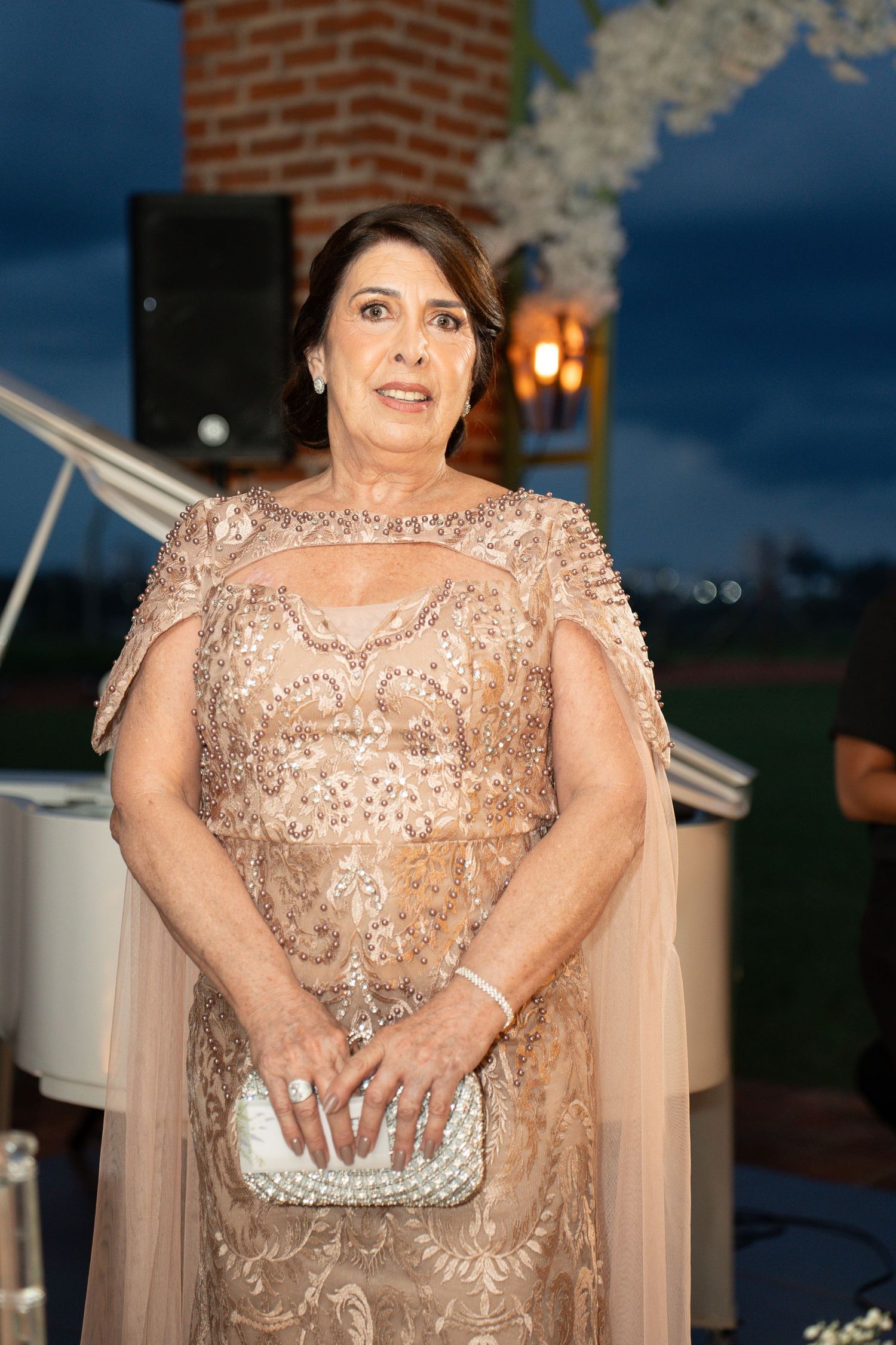 Elegant older woman in gold beaded gown holding white clutch at formal outdoor evening event.