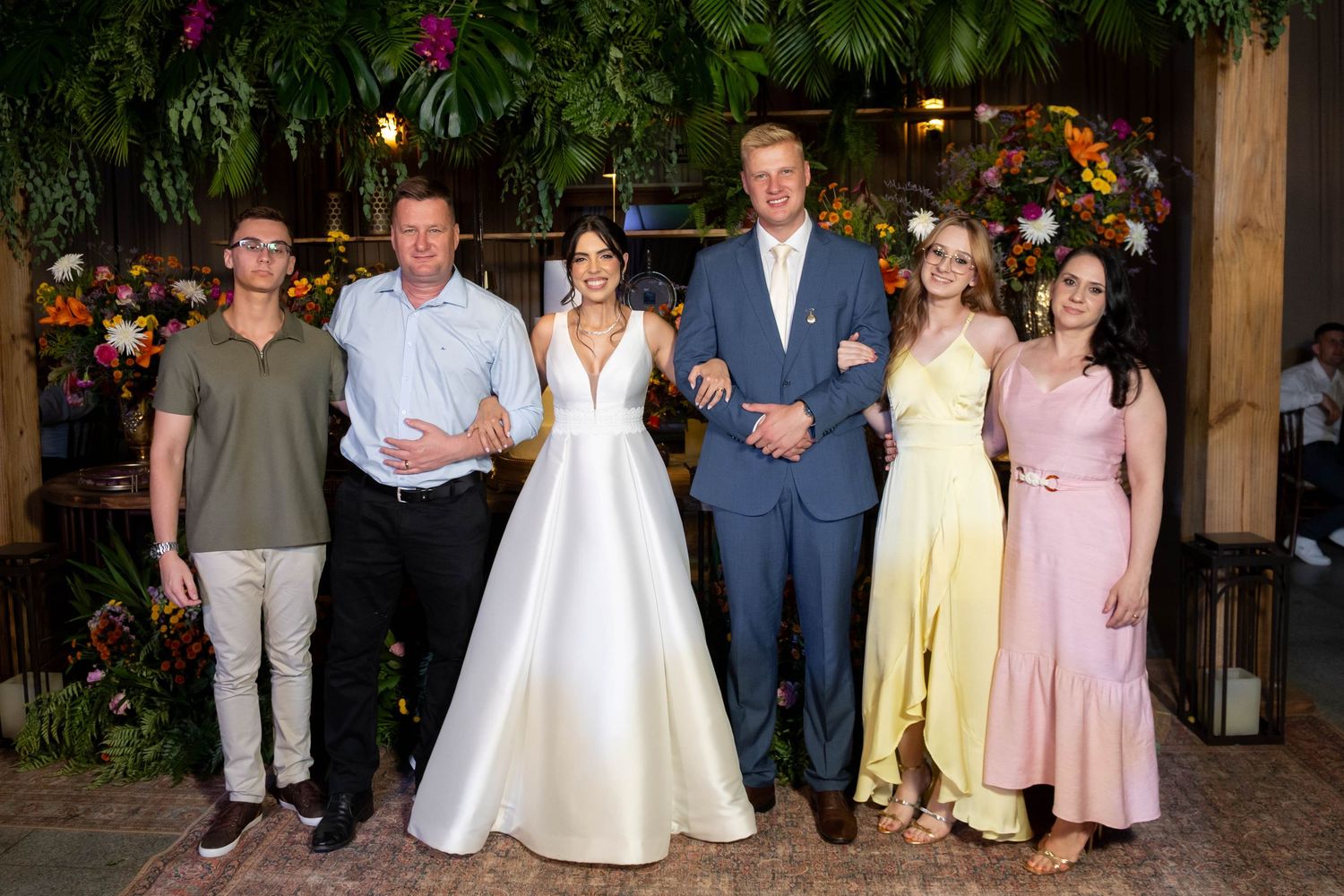 A joyful wedding party of six poses together indoors, surrounded by lush tropical greenery and colorful floral arrangements.
