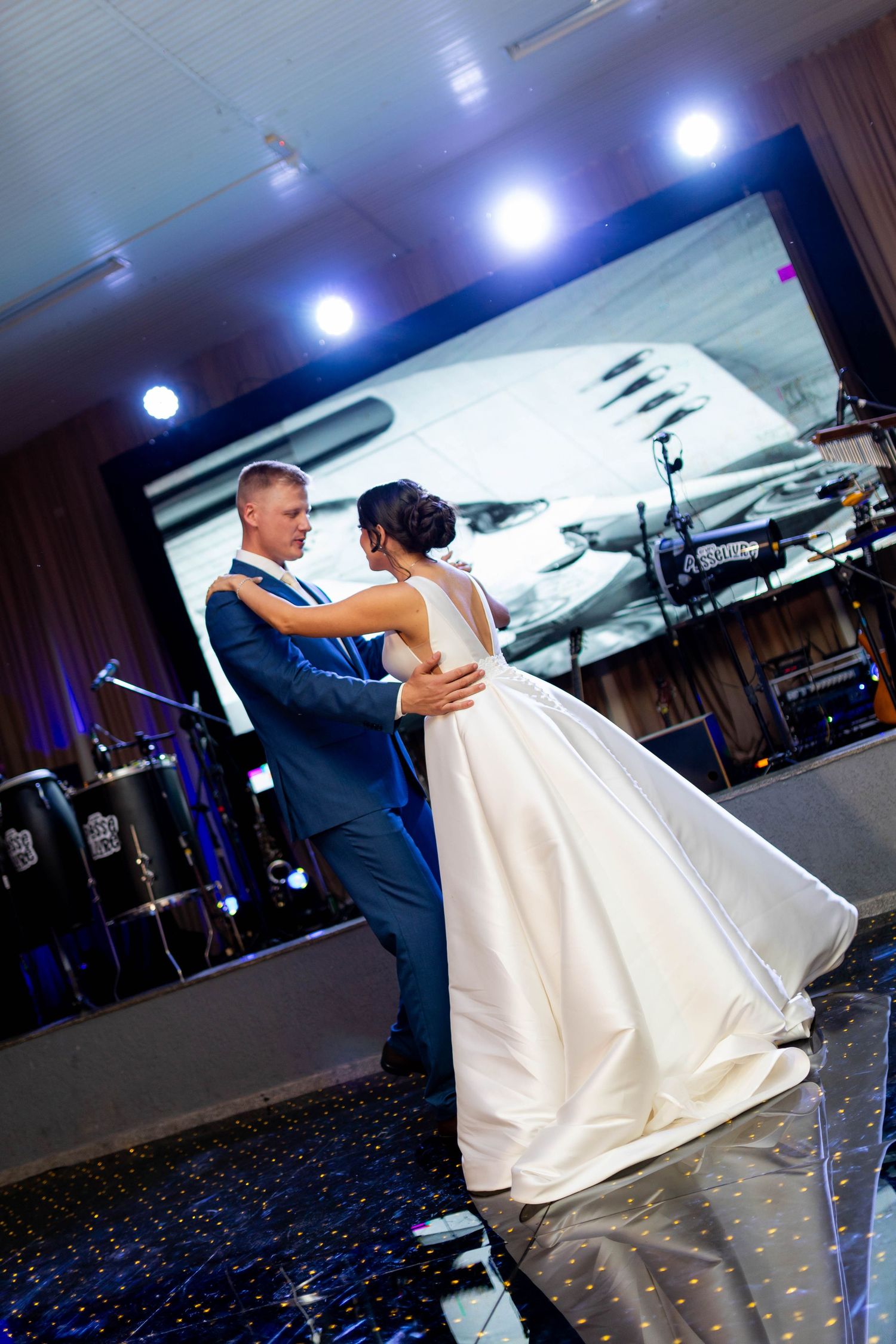 Bride and groom share their first dance at a wedding reception with stage lights and a large screen backdrop.