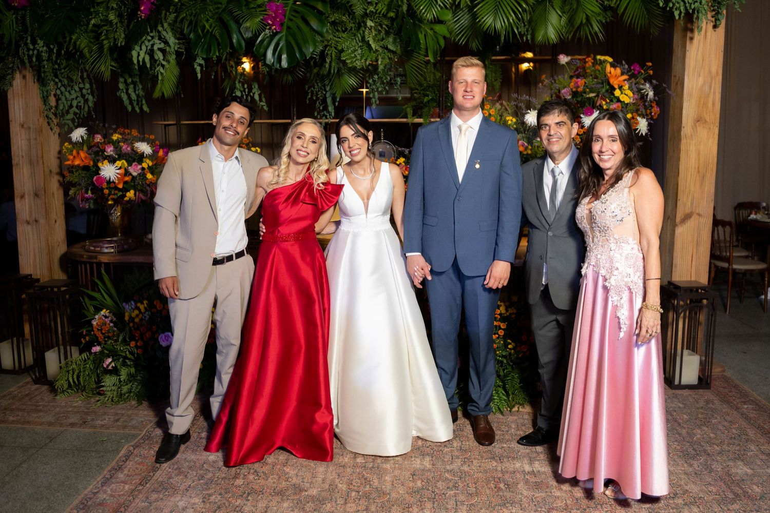 A wedding party of six poses elegantly in formal attire, including a bride in white and guests in red and pink gowns.