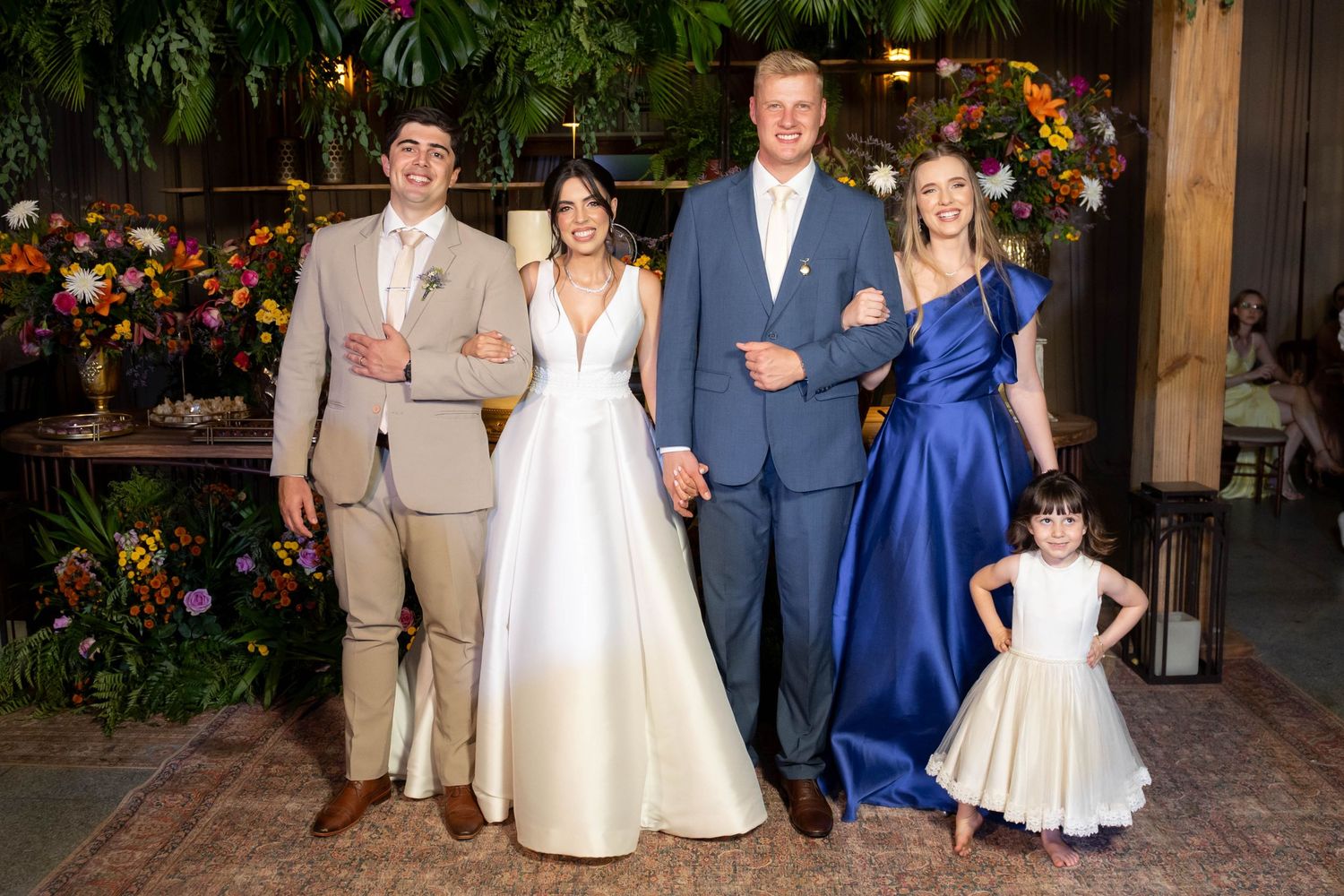 Wedding party poses together, featuring bride in white gown, groom, guests in blue and tan formal wear, and flower girl.
