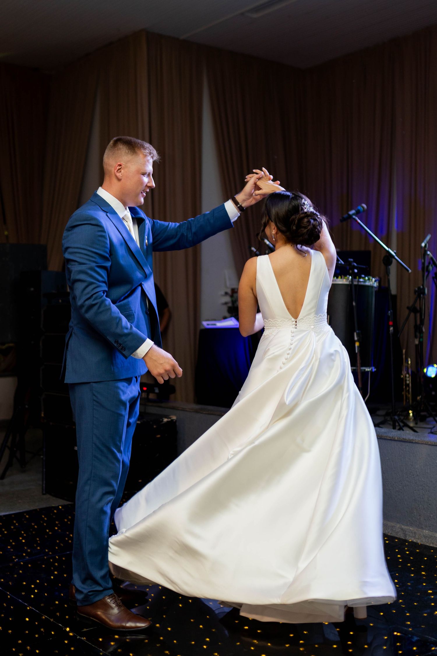 Bride and groom share their first dance, groom in blue suit twirling bride in white gown on starry dance floor.
