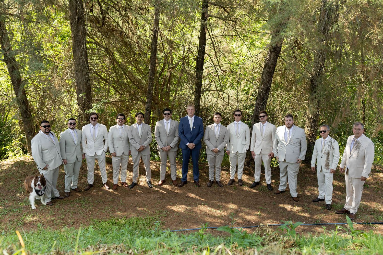 Groom in blue suit stands with groomsmen in tan suits and sunglasses outdoors in a lush forest wedding photo.