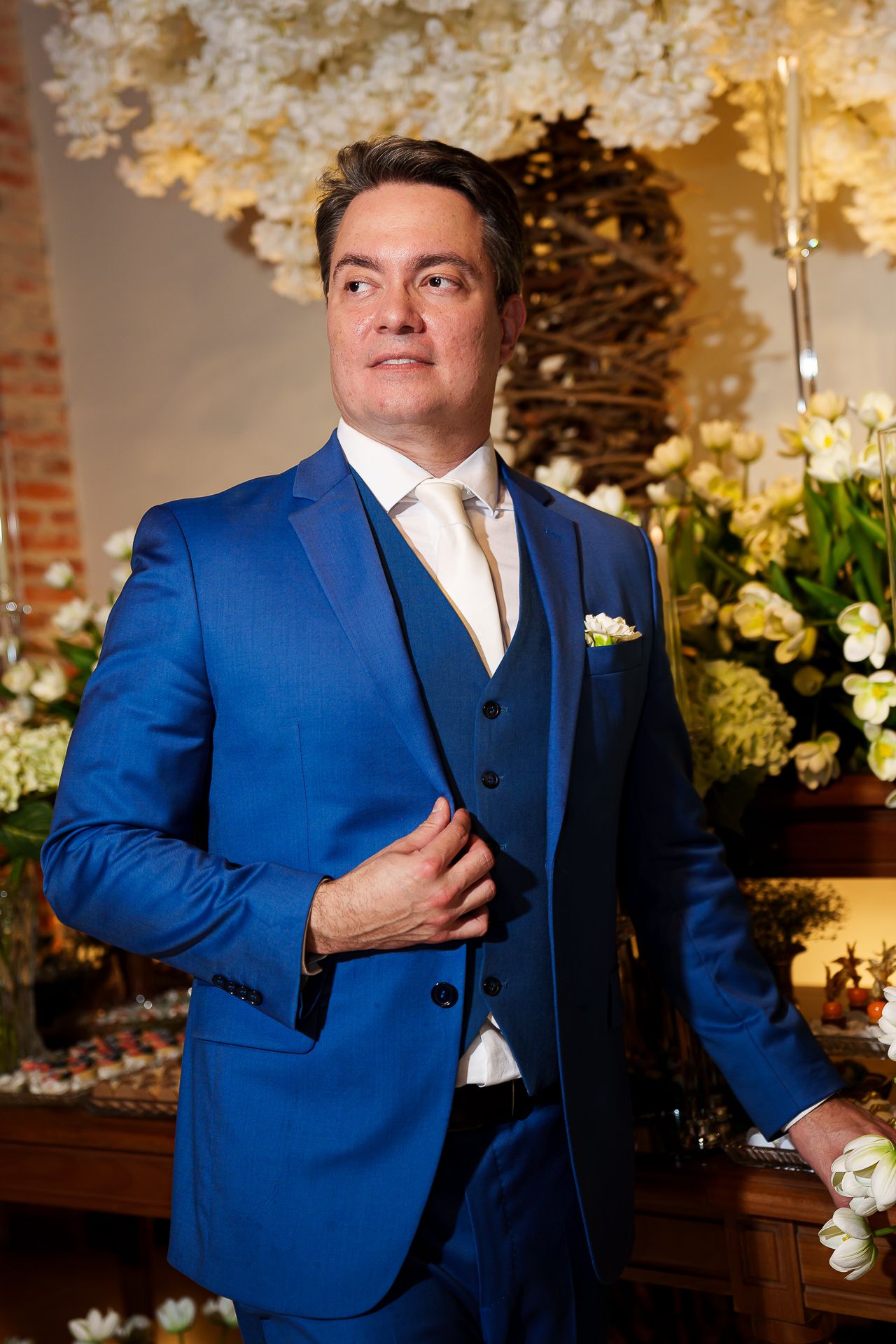 Elegant man in blue three-piece suit with white boutonniere poses before floral wedding backdrop.