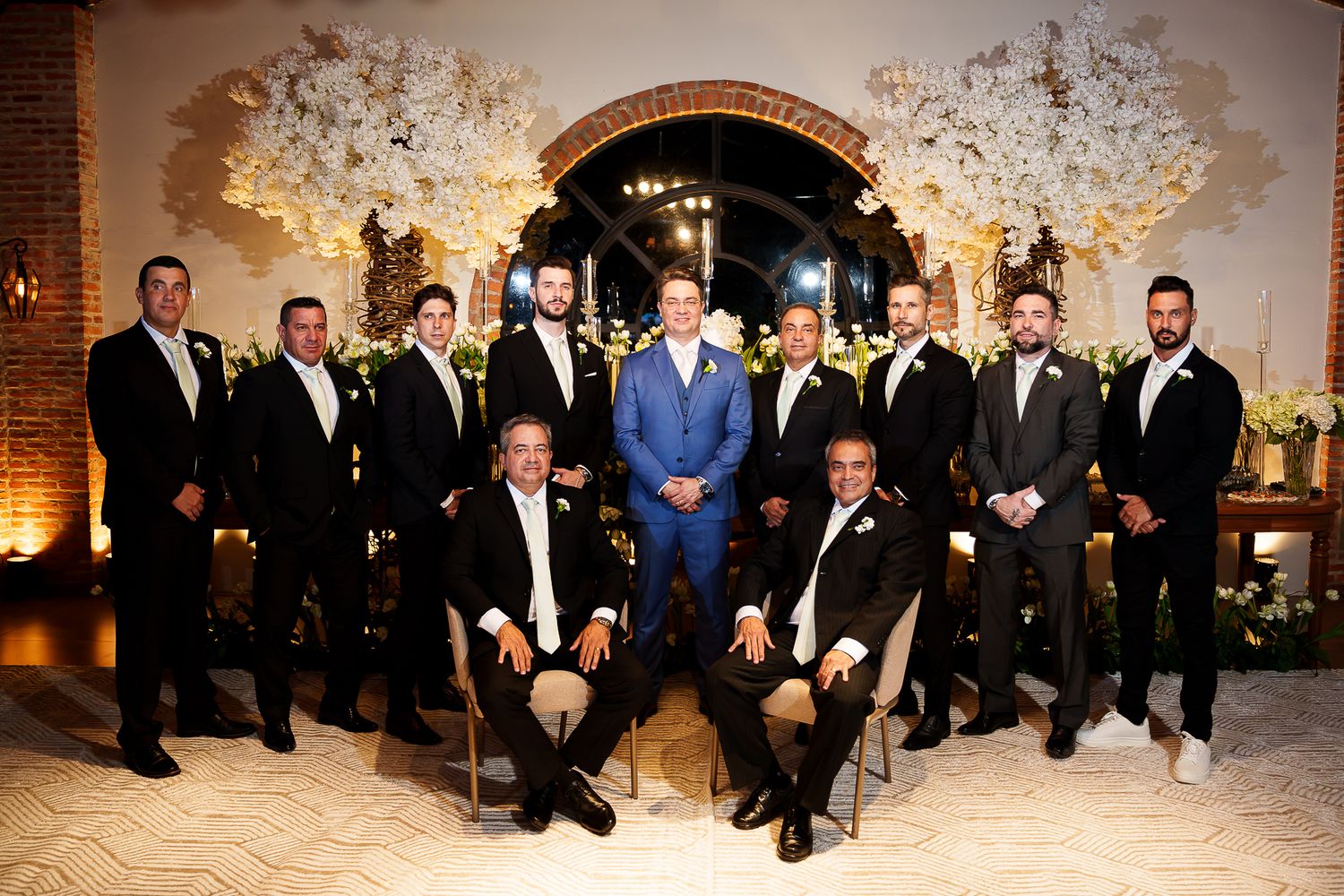 Groom in blue suit poses with groomsmen in black tuxedos before floral backdrop at elegant wedding venue.