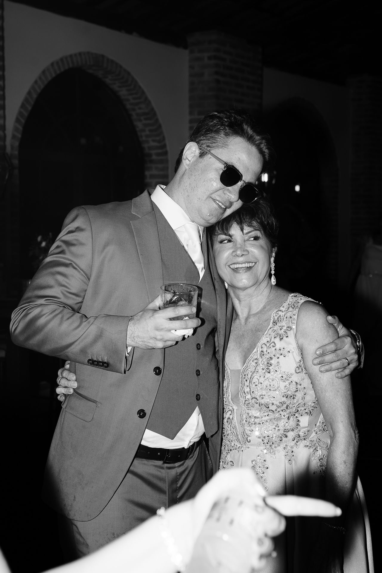 Black and white photo of a couple posing together at a formal event, man in suit with sunglasses holding a drink.