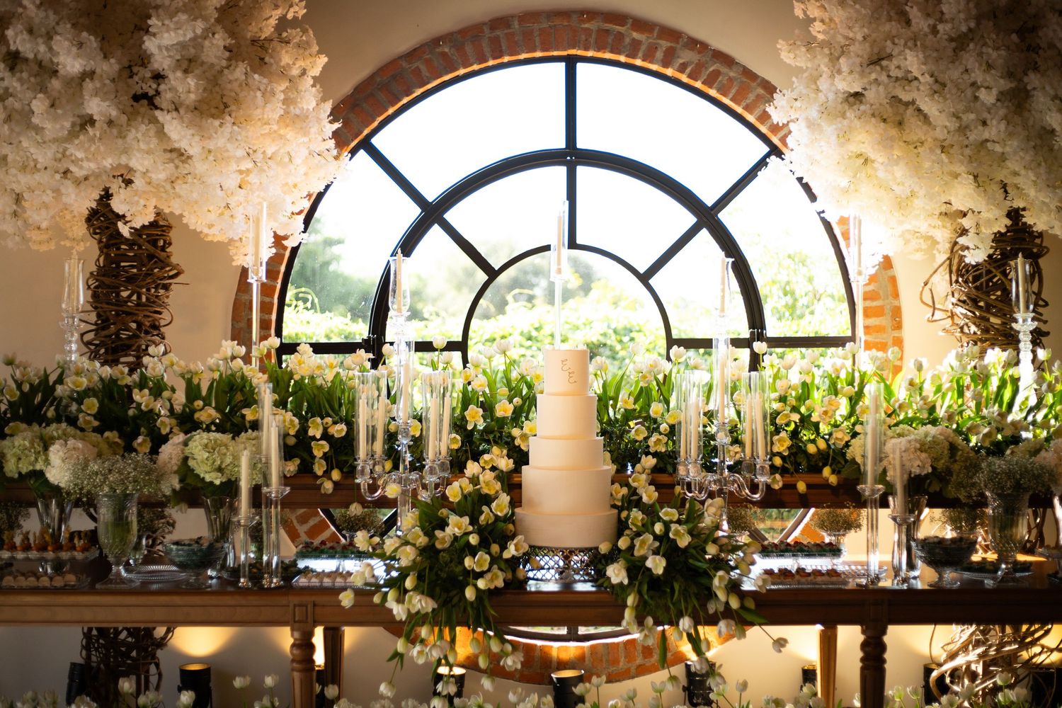 Elegant wedding cake display with white floral arch, candles, and lush arrangements beneath a grand arched window.