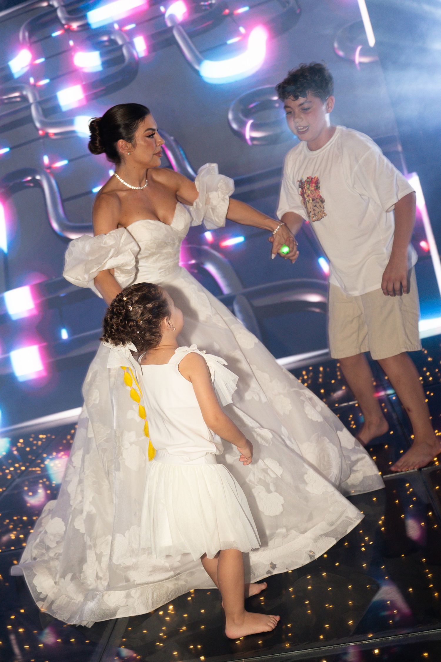 Bride in white gown dances with two children on a glowing dance floor with colorful lights.