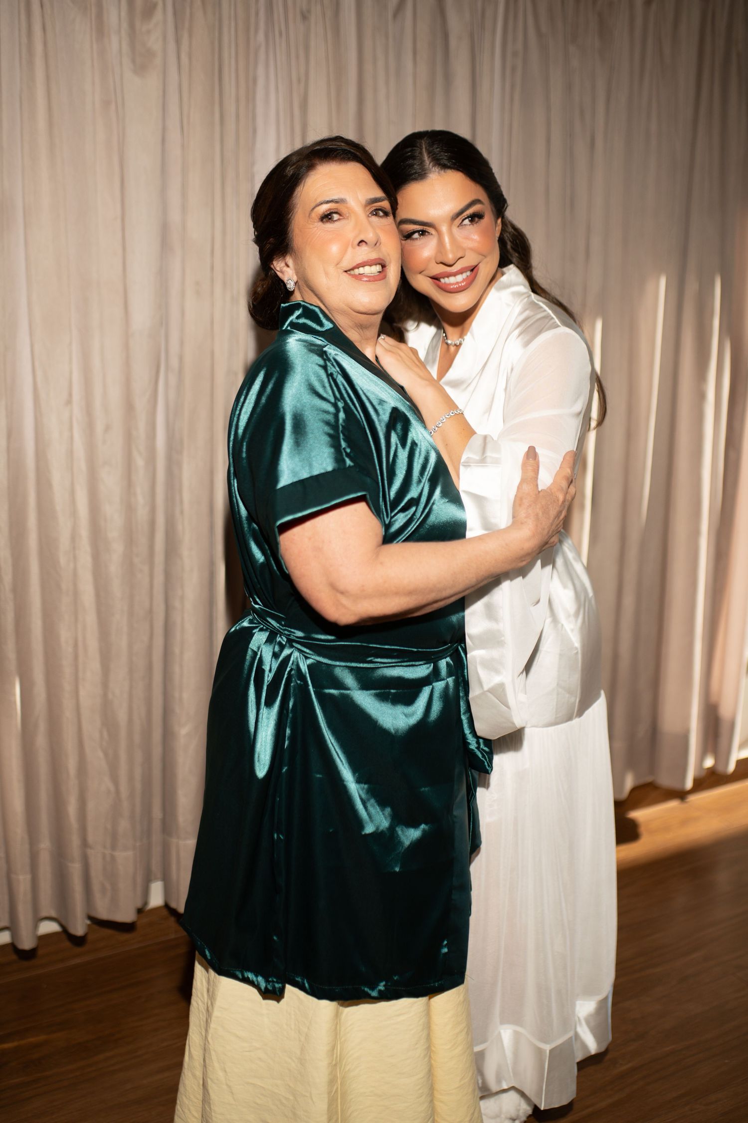Two women embracing and smiling, one in a teal satin wrap dress, the other in a white outfit, posing together indoors.