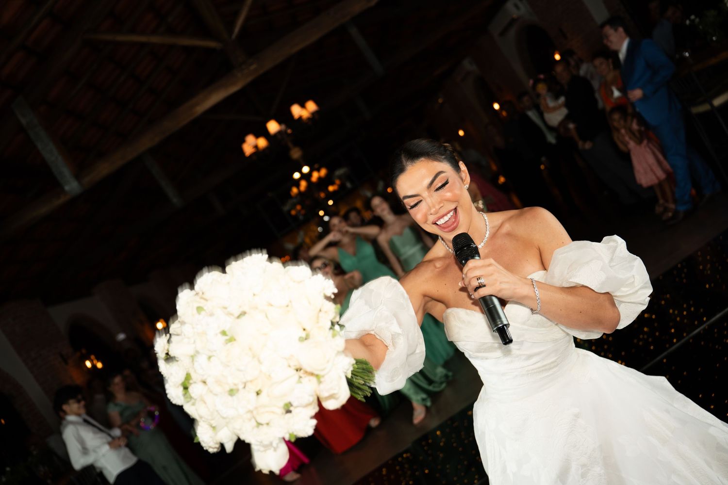 Bride in white wedding dress holding bouquet of white roses while speaking into microphone at wedding reception.