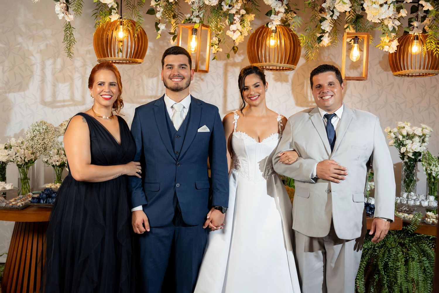 A wedding party of four poses elegantly at a floral-decorated reception venue with golden pendant lights overhead.