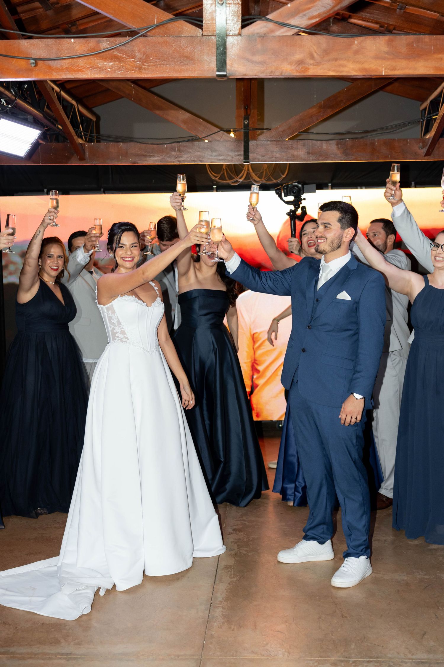 Bride and groom raise champagne glasses with wedding party in navy attire during indoor reception celebration.