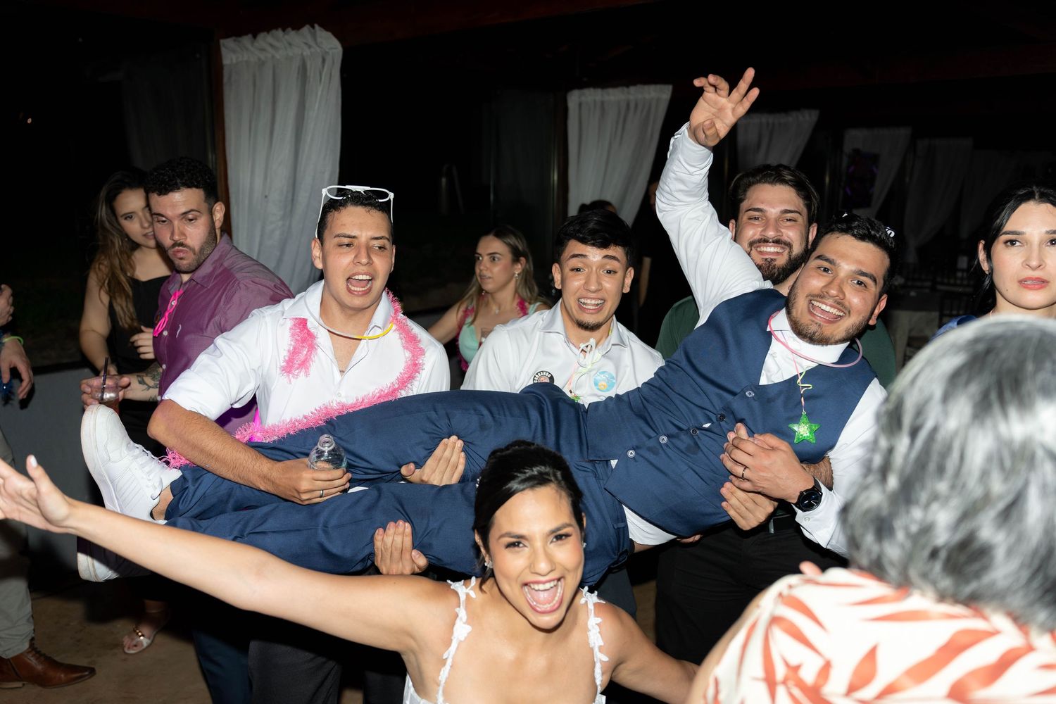 Bride in white dress lifted by groomsmen in navy suits celebrating joyfully at a wedding reception party.