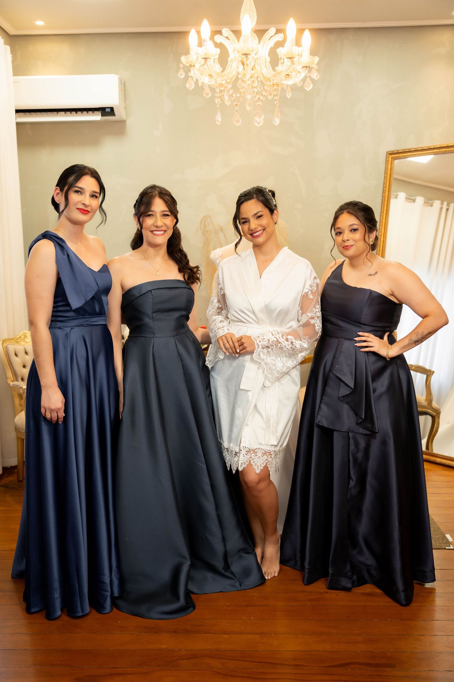 Bride in white lace robe poses with three bridesmaids in navy blue gowns under a crystal chandelier.