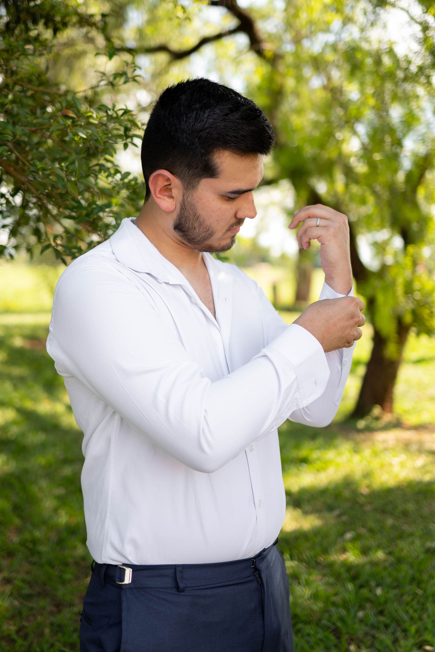 A young man in a white dress shirt adjusts his cufflinks outdoors in a lush green park setting.