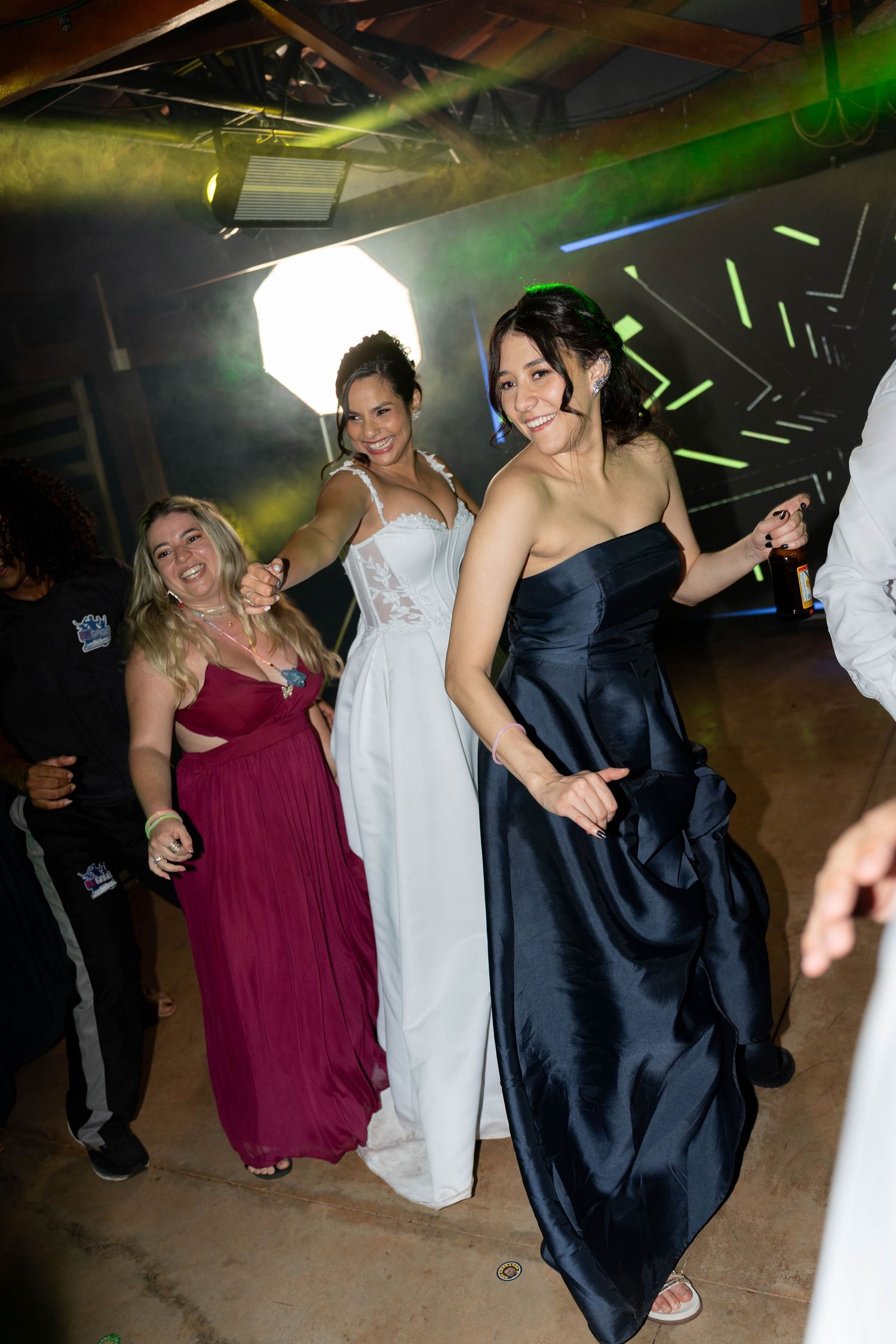 Three women in formal gowns dancing at a wedding reception under colorful lights.
