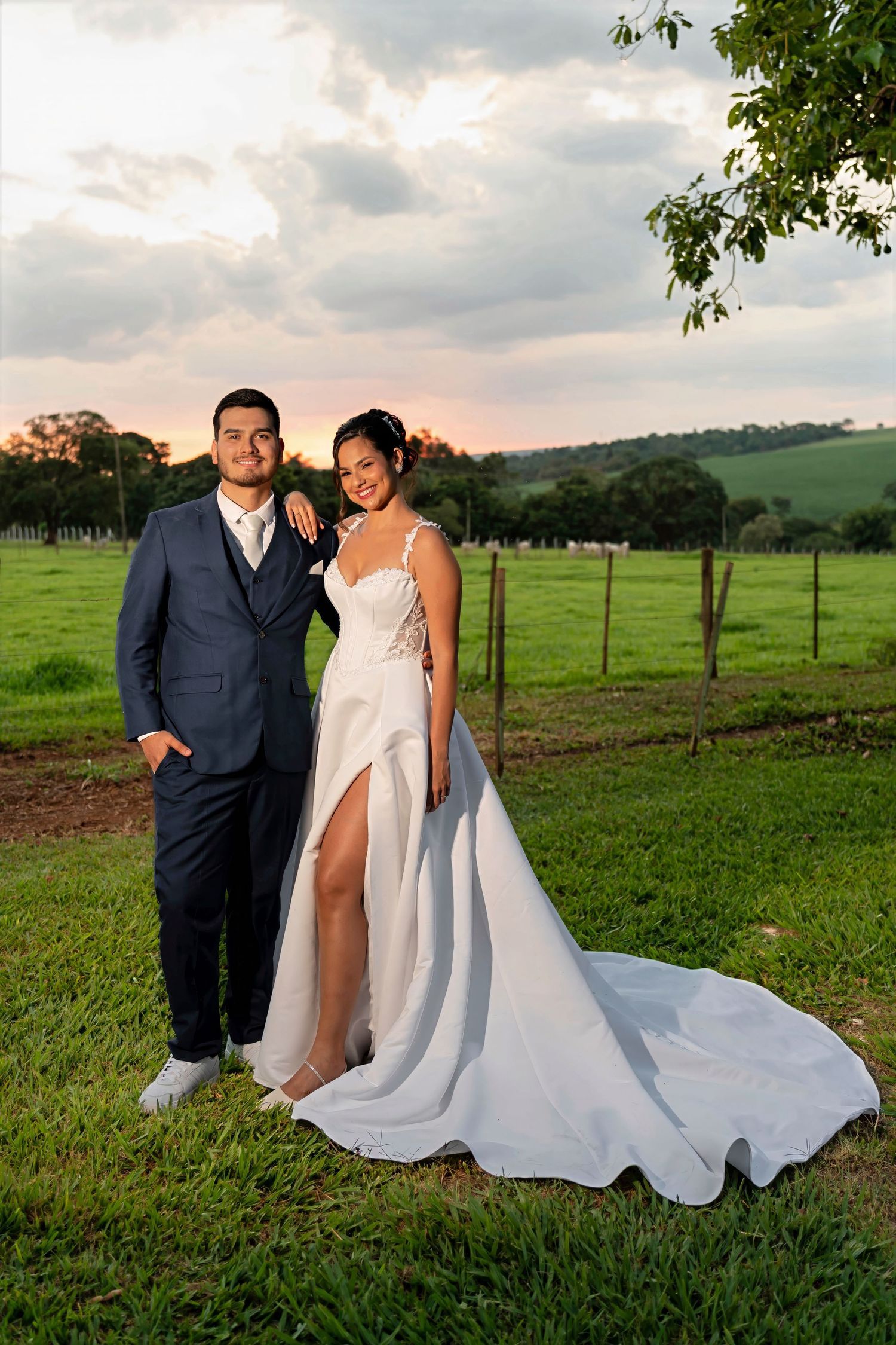 Bride and groom posing in a lush green field at sunset, bride in white gown with train, groom in navy suit.