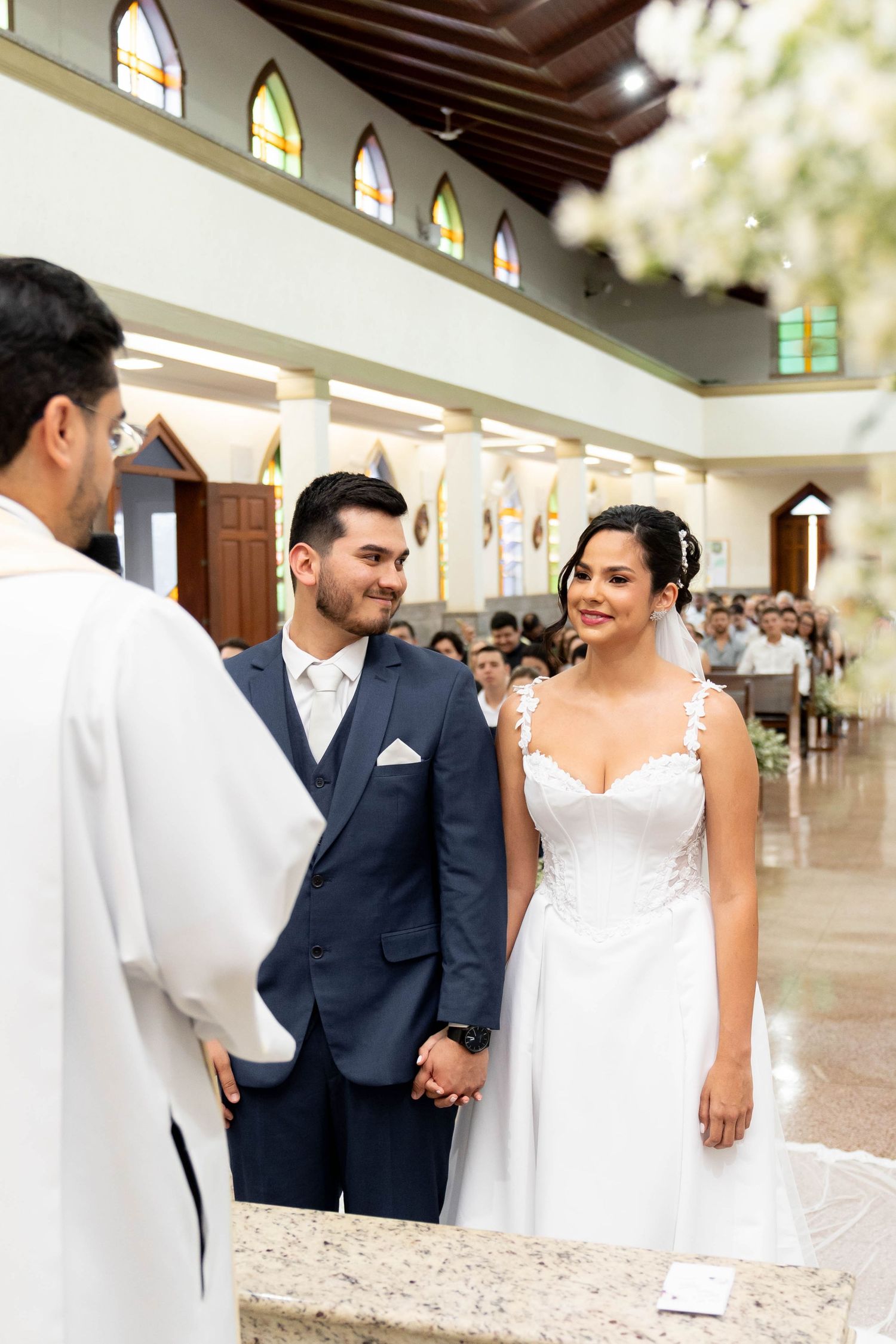 Bride and groom hold hands at church altar during wedding ceremony, smiling at officiant in white robes.
