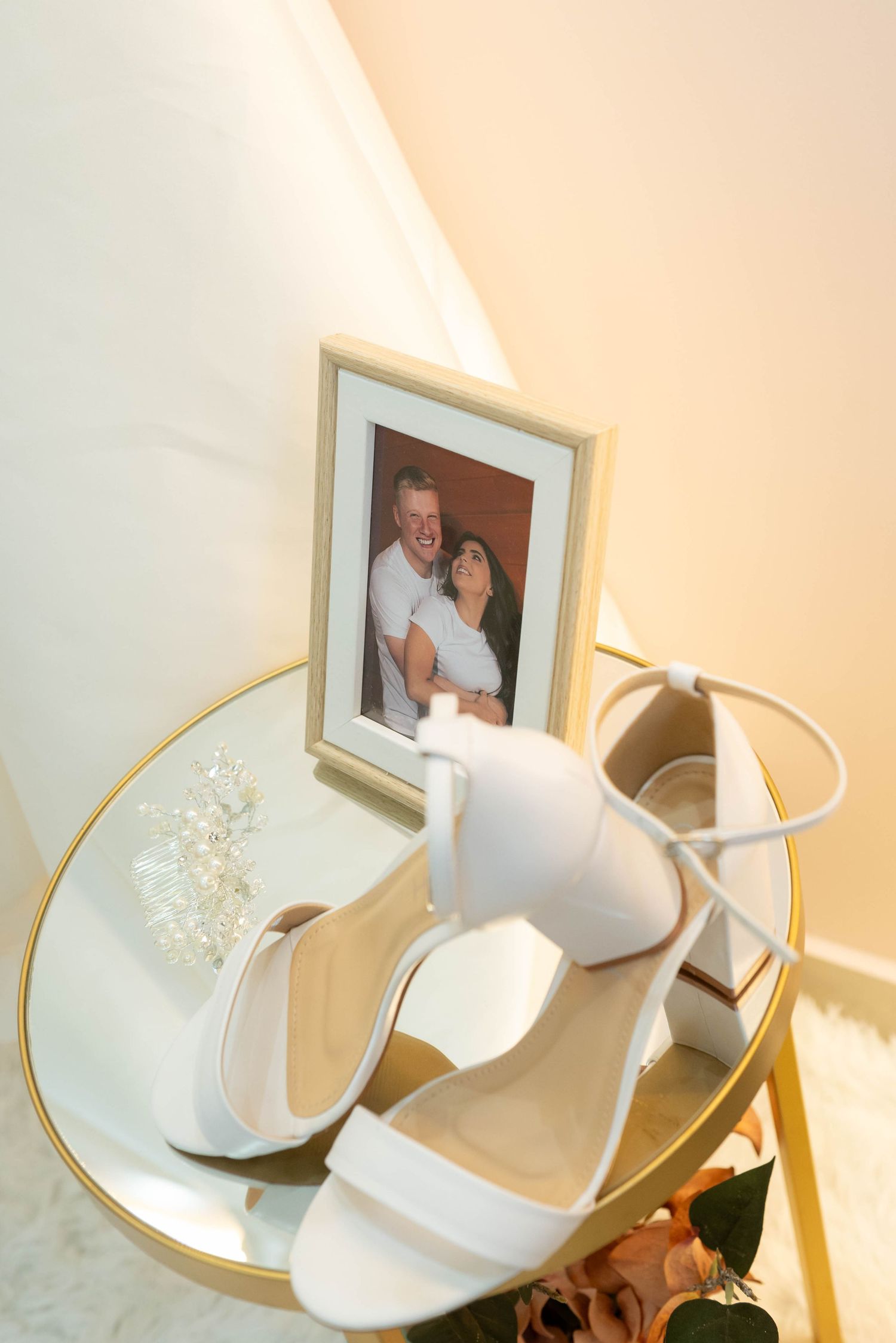 White bridal heels displayed on a glass table beside a framed couple photo and pearl hair accessories.