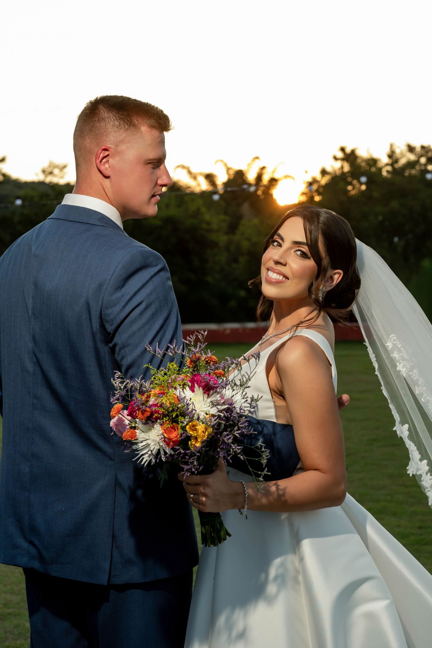 Bride smiling at groom in blue suit at sunset, holding colorful bouquet in white gown with veil outdoors.