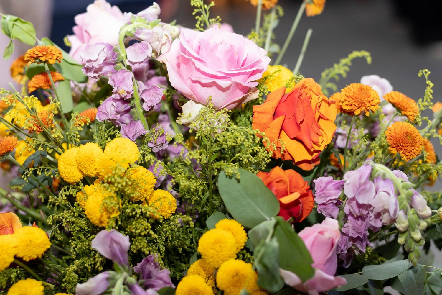 Vibrant bouquet featuring pink roses, orange blooms, yellow marigolds, and purple flowers with lush green foliage.