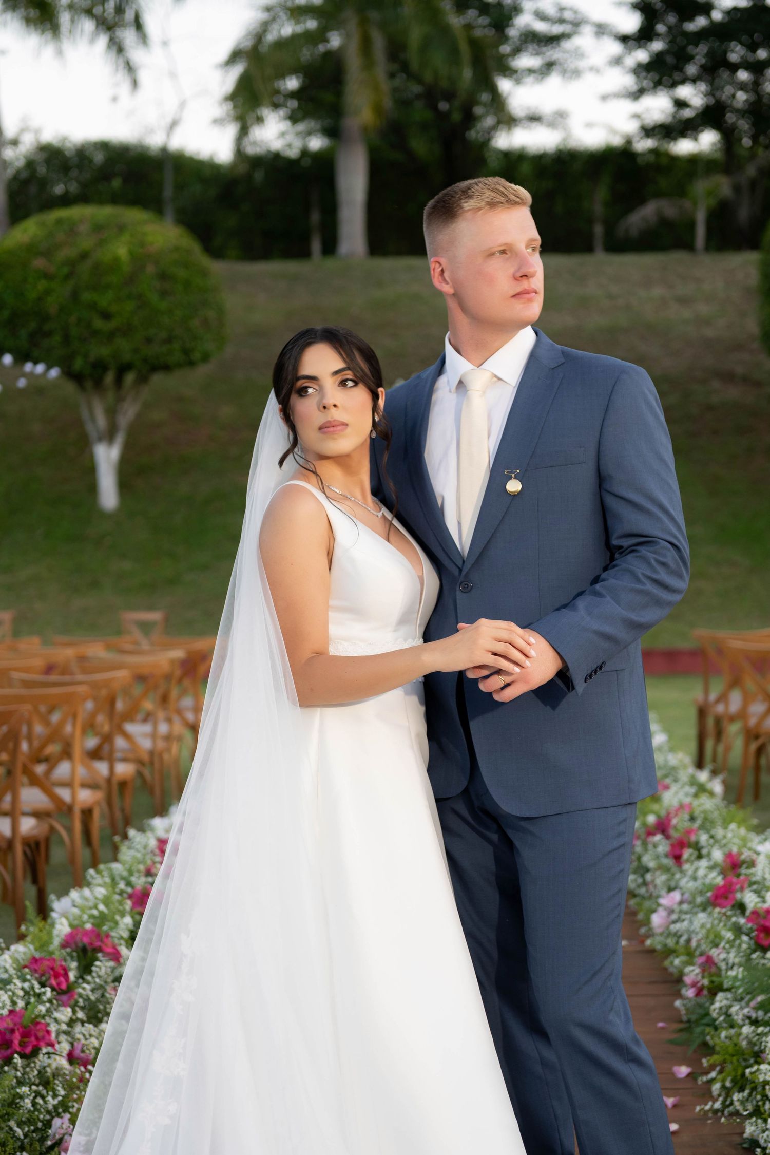 Bride in white gown and groom in blue suit pose together at outdoor wedding ceremony with floral aisle.