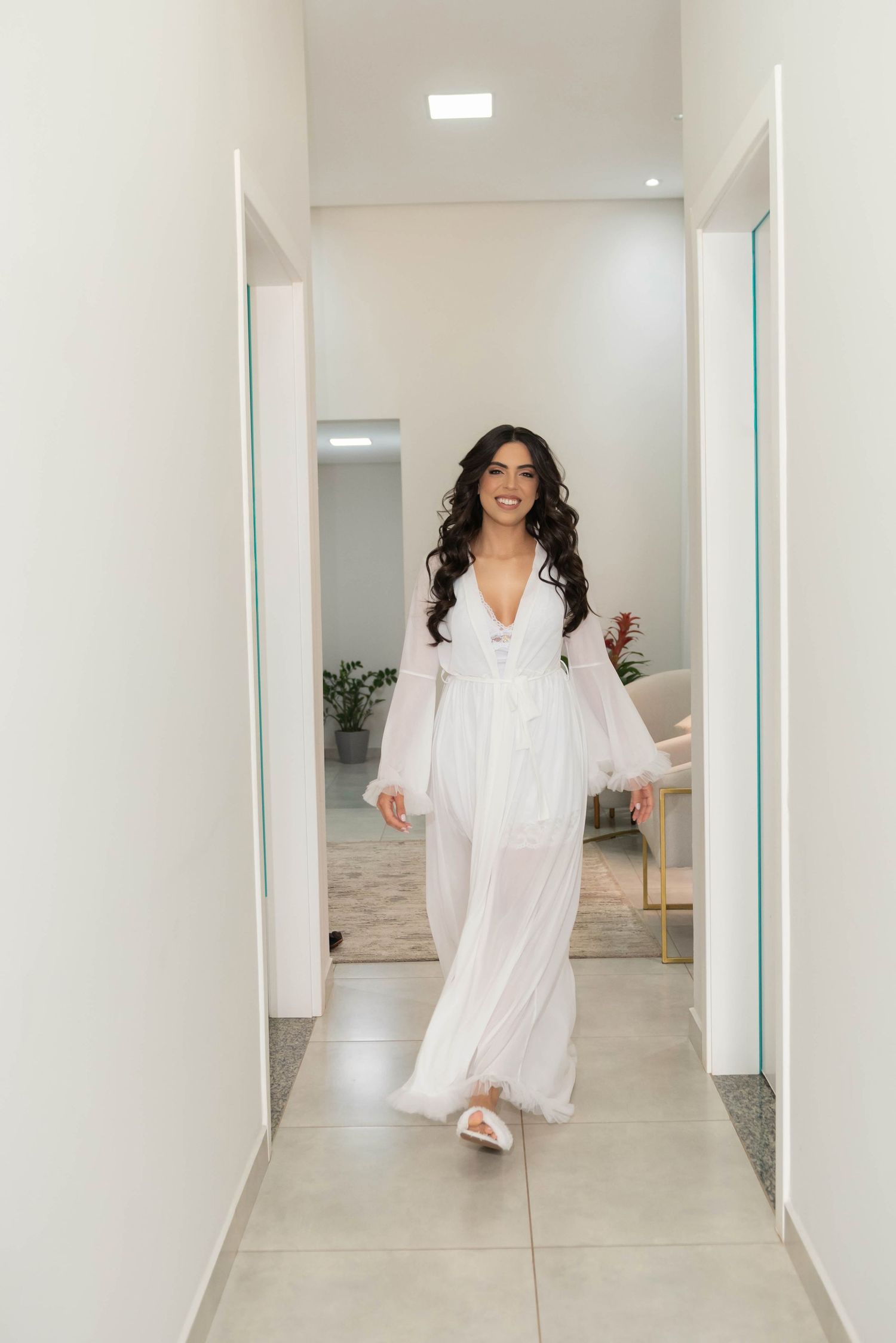 Smiling bride in white sheer robe walking down a bright modern hallway on her wedding day.