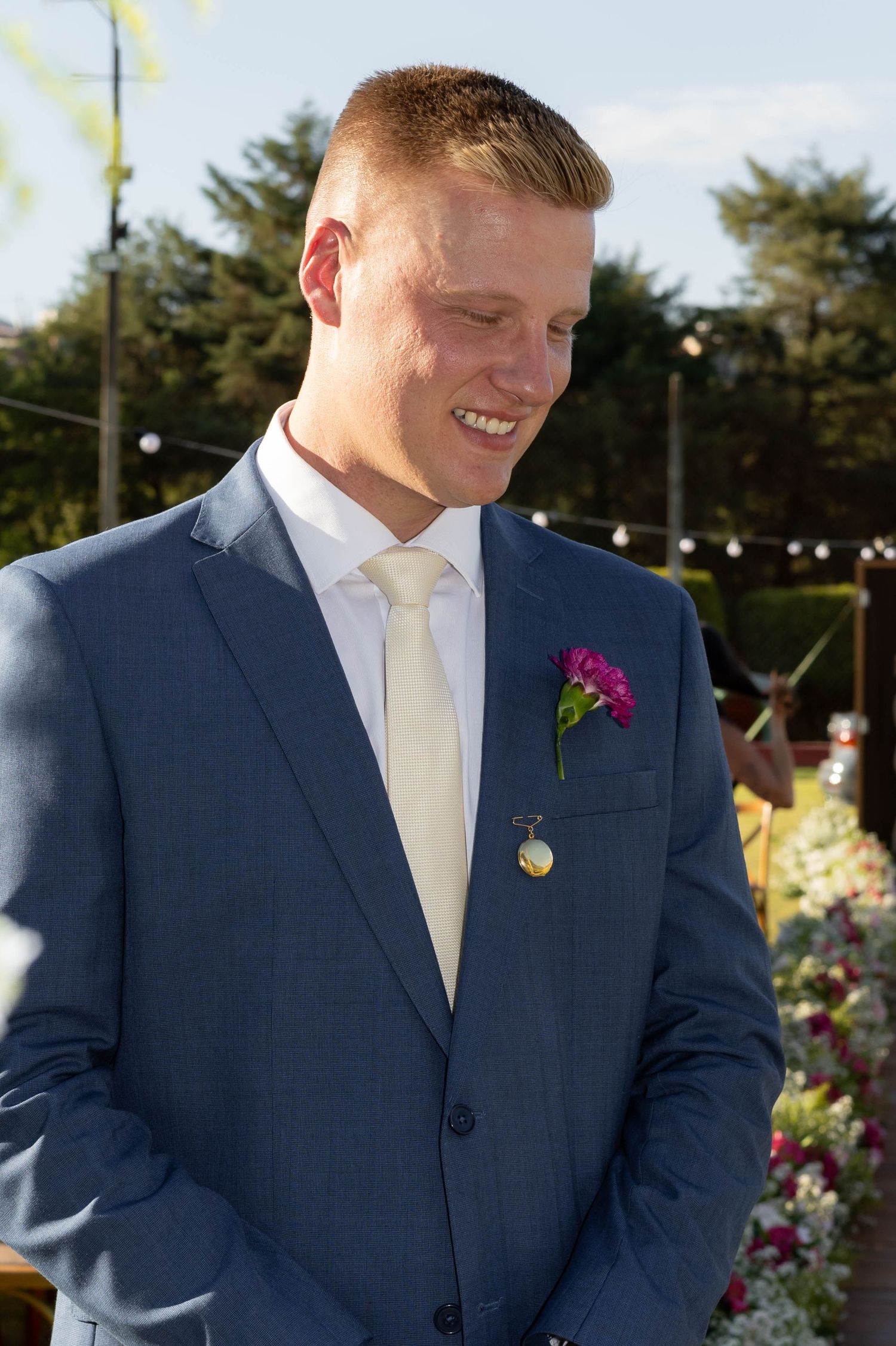 Smiling groom in blue suit with pink boutonniere and cream tie at outdoor wedding ceremony.