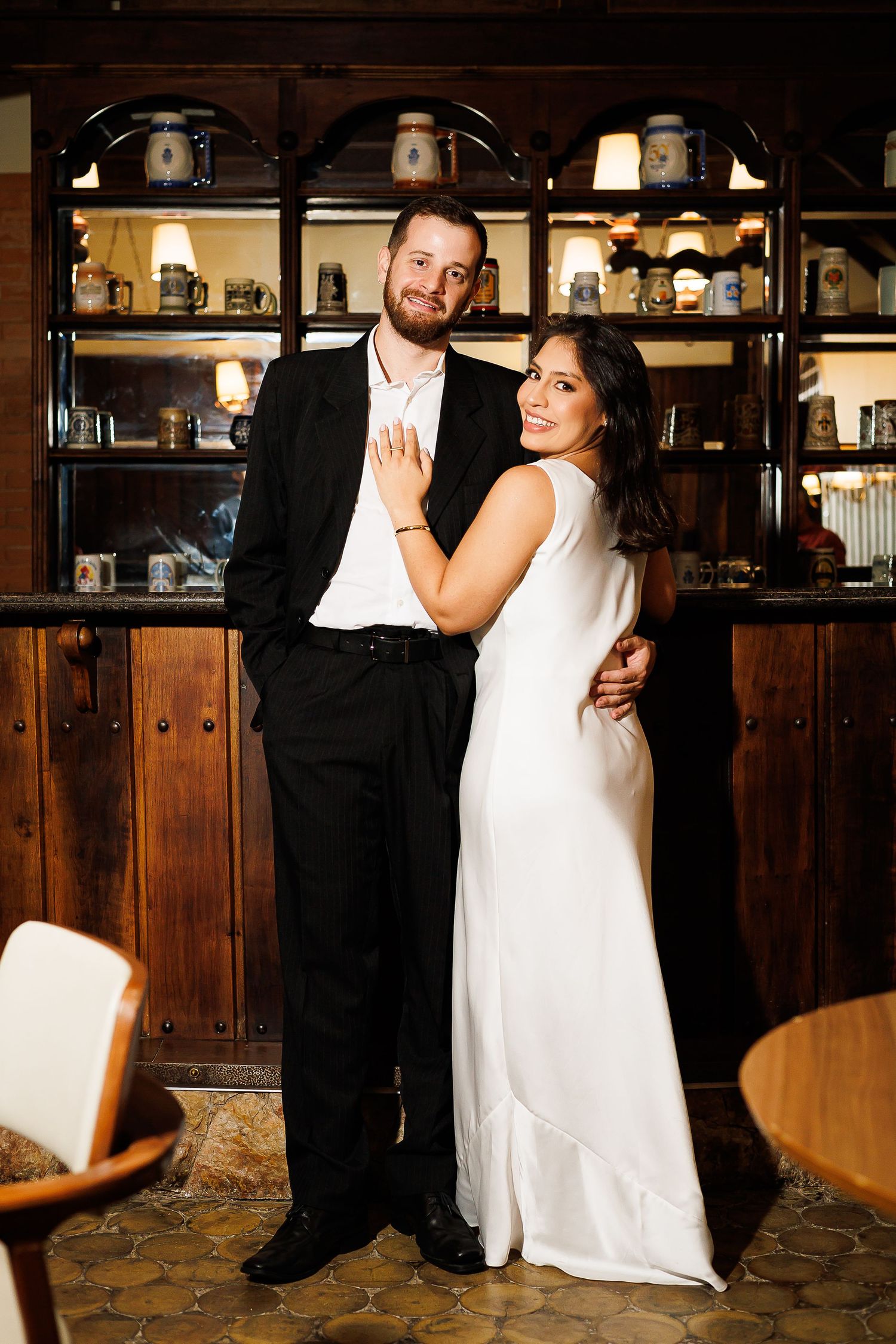 A couple in wedding attire poses together in a warmly lit restaurant, the bride in white and groom in black suit.