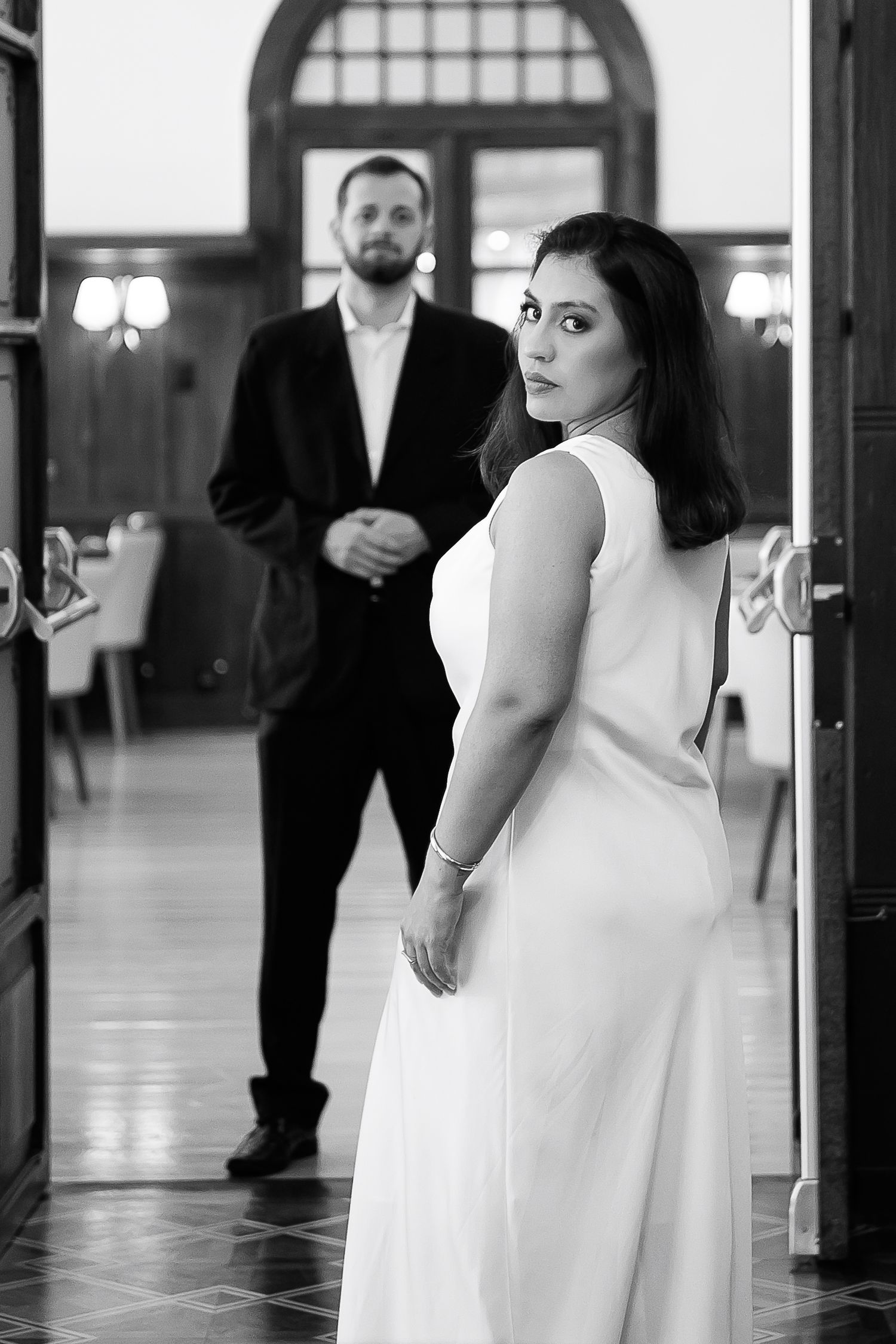 Black and white wedding photo of bride in white dress looking back at groom standing in elegant hall.