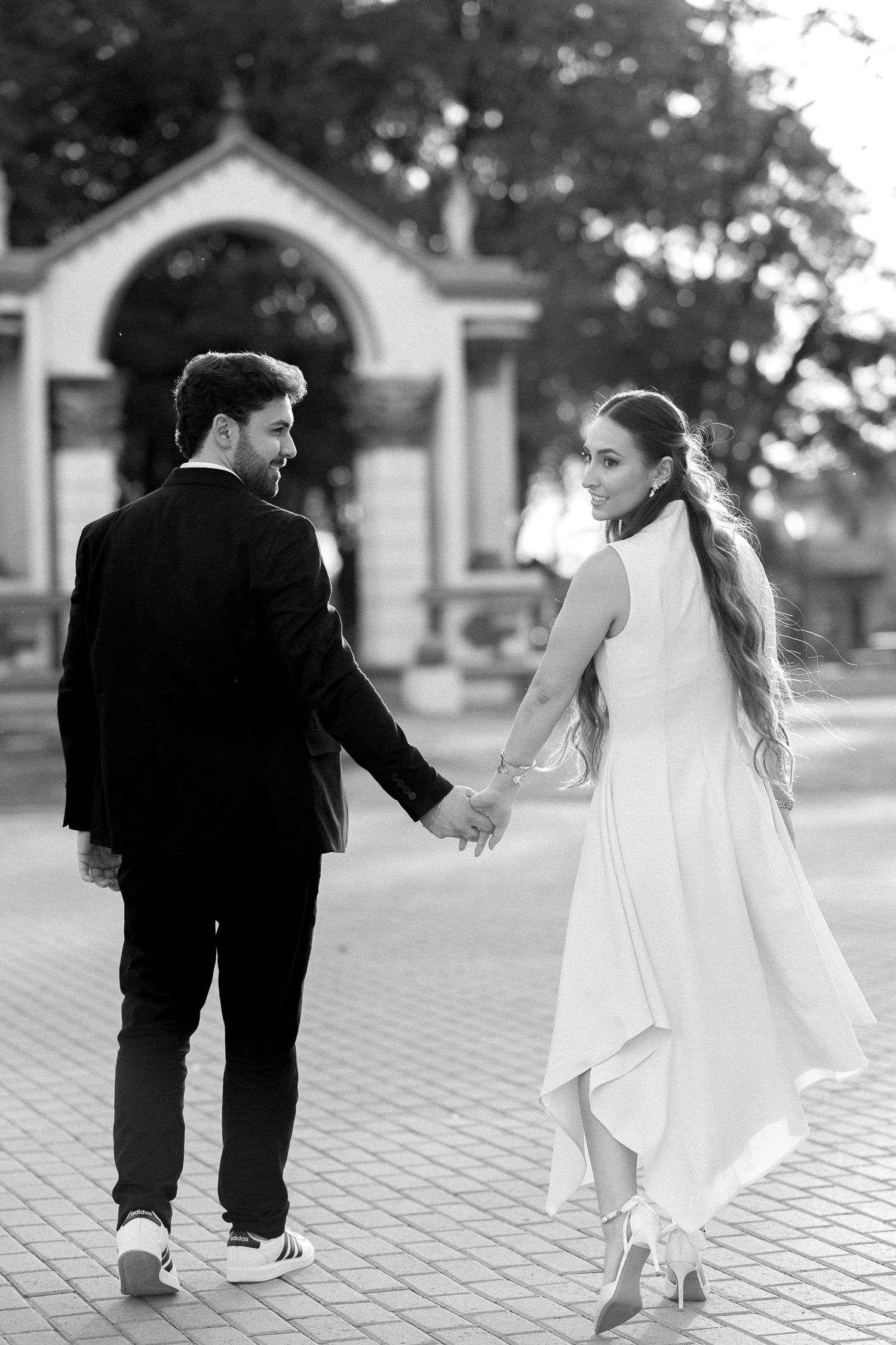 Black and white photo of a bride and groom holding hands, walking away from camera in elegant wedding attire.