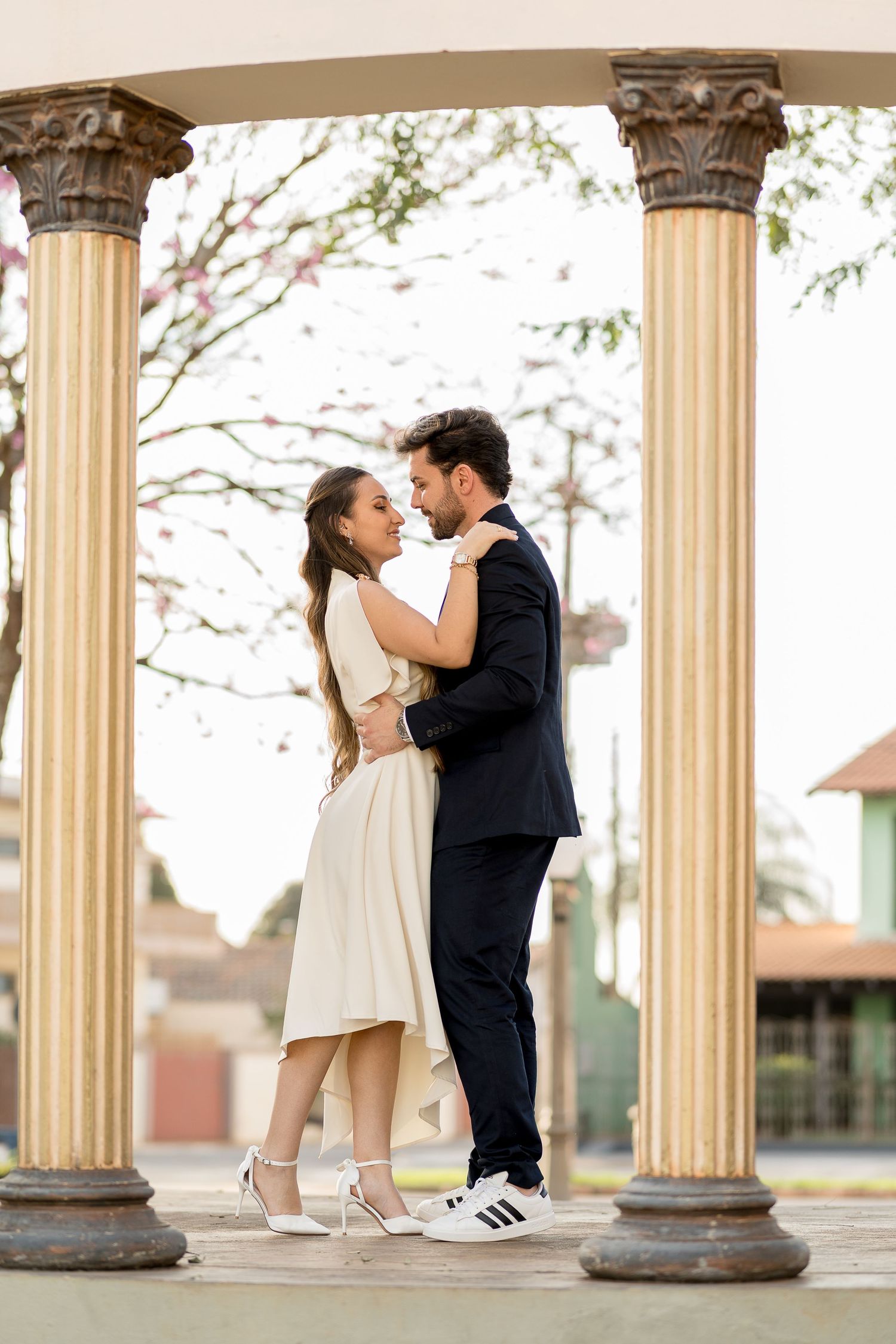 Couple embracing romantically between elegant columns in an outdoor plaza, woman in white dress and man in dark suit.