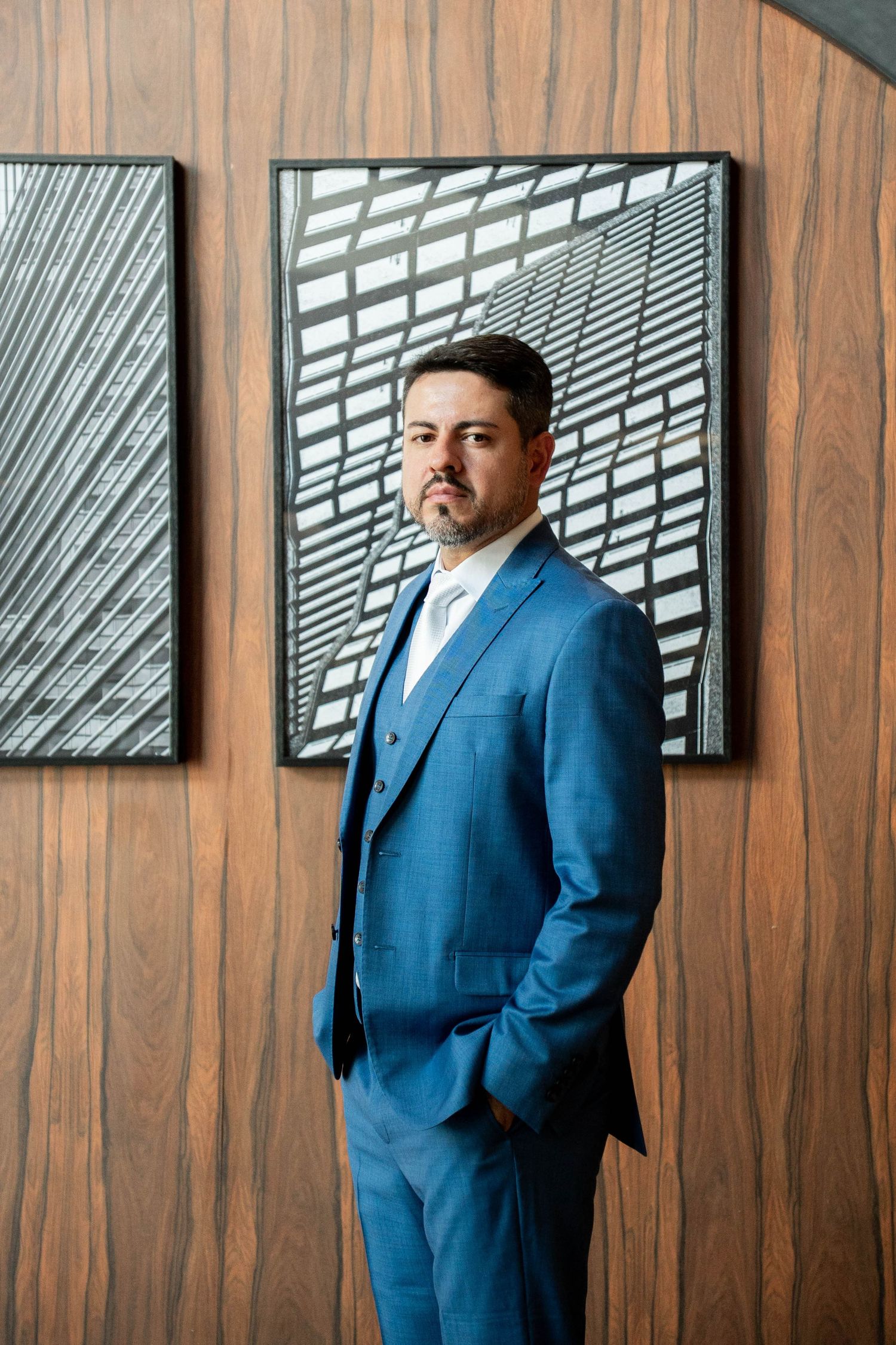 A confident man in a blue three-piece suit stands before a wooden wall with framed architectural photography.
