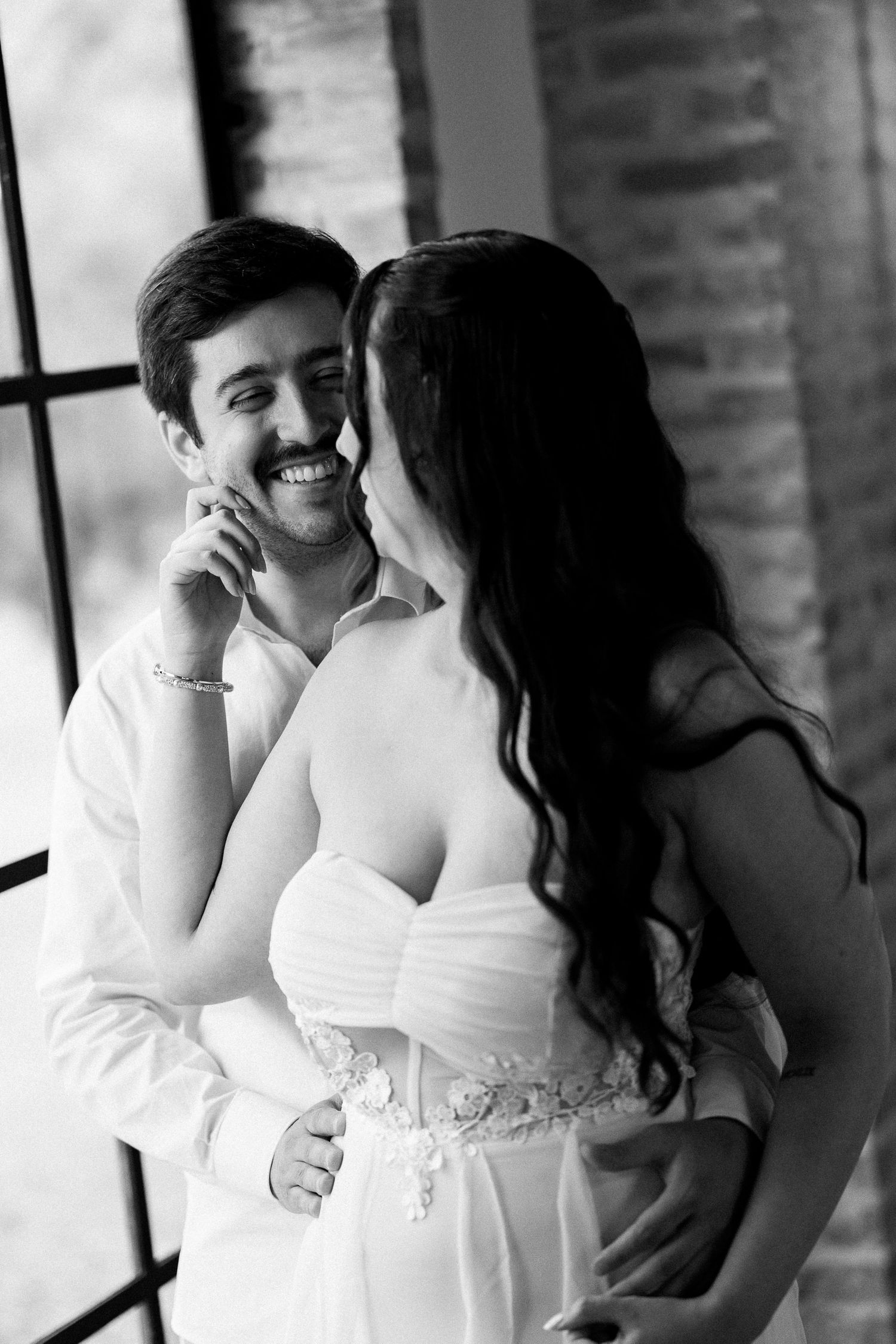 Black and white photo of smiling groom and bride in white dress sharing a tender moment near a window.