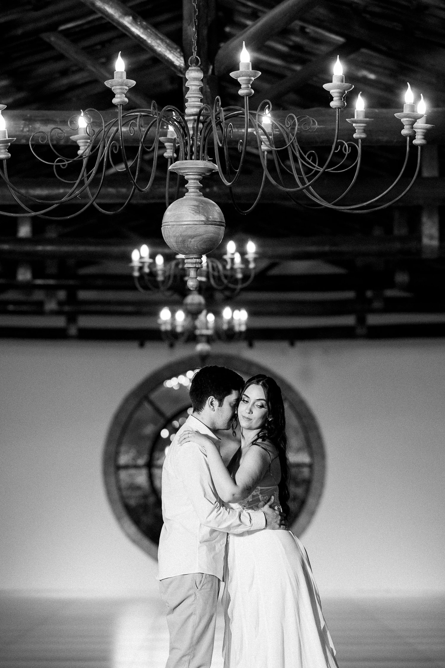 Black and white photo of a couple sharing a romantic dance under elegant chandeliers in a rustic venue.
