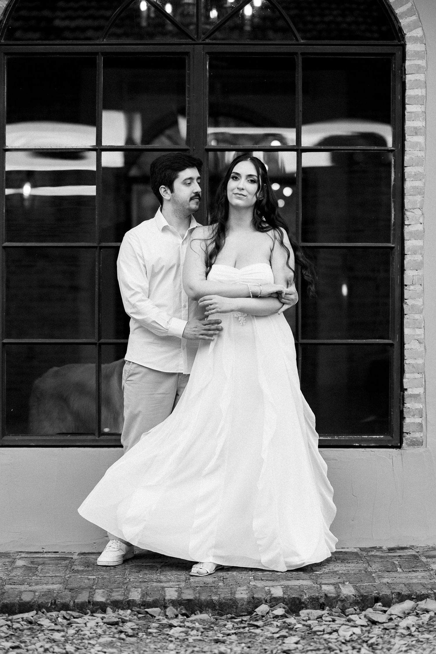 Black and white photo of a couple posing outdoors, woman in flowing white gown, man in casual shirt embracing her.