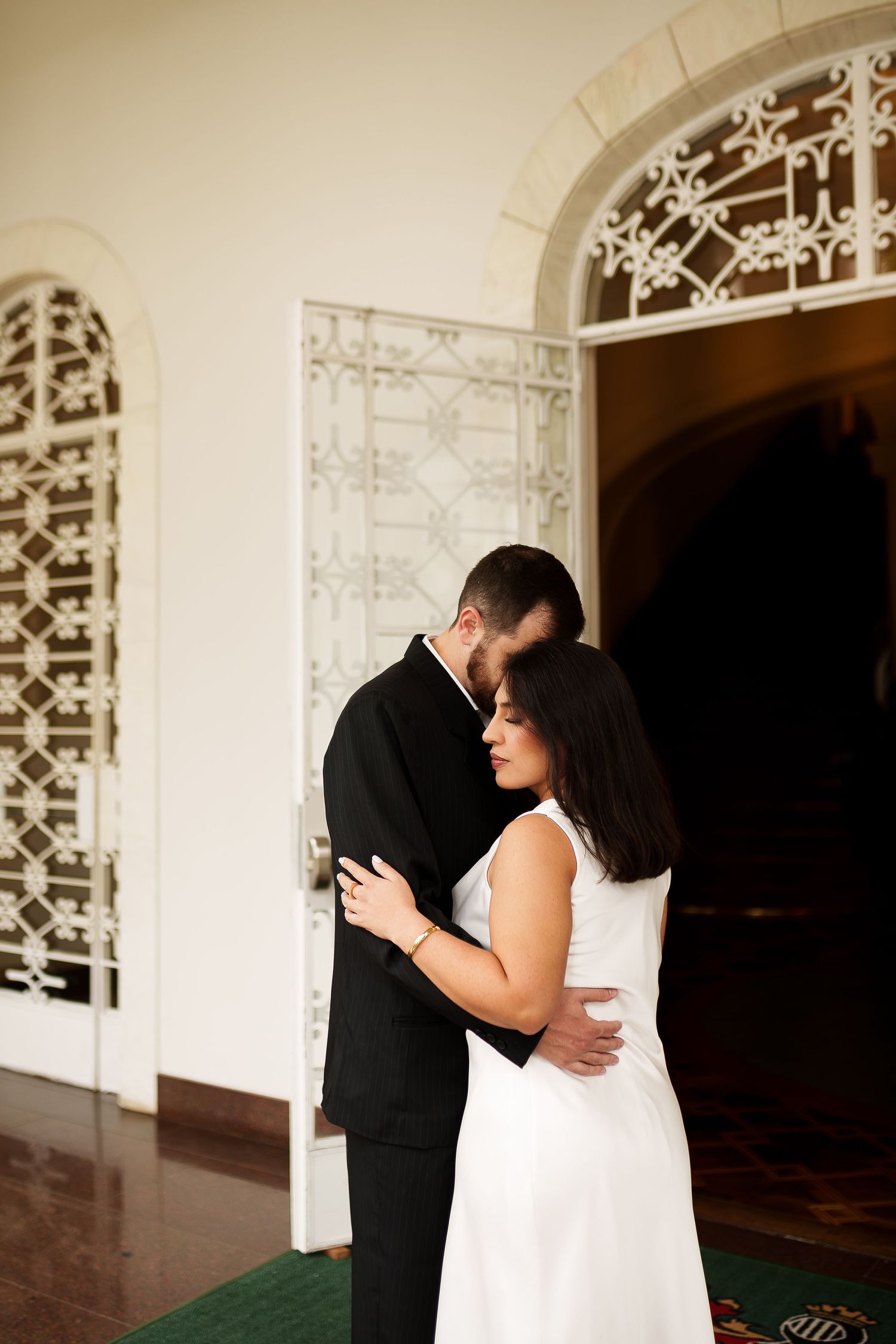 A couple in formal attire embraces tenderly near an ornate white arched doorway in an elegant venue.