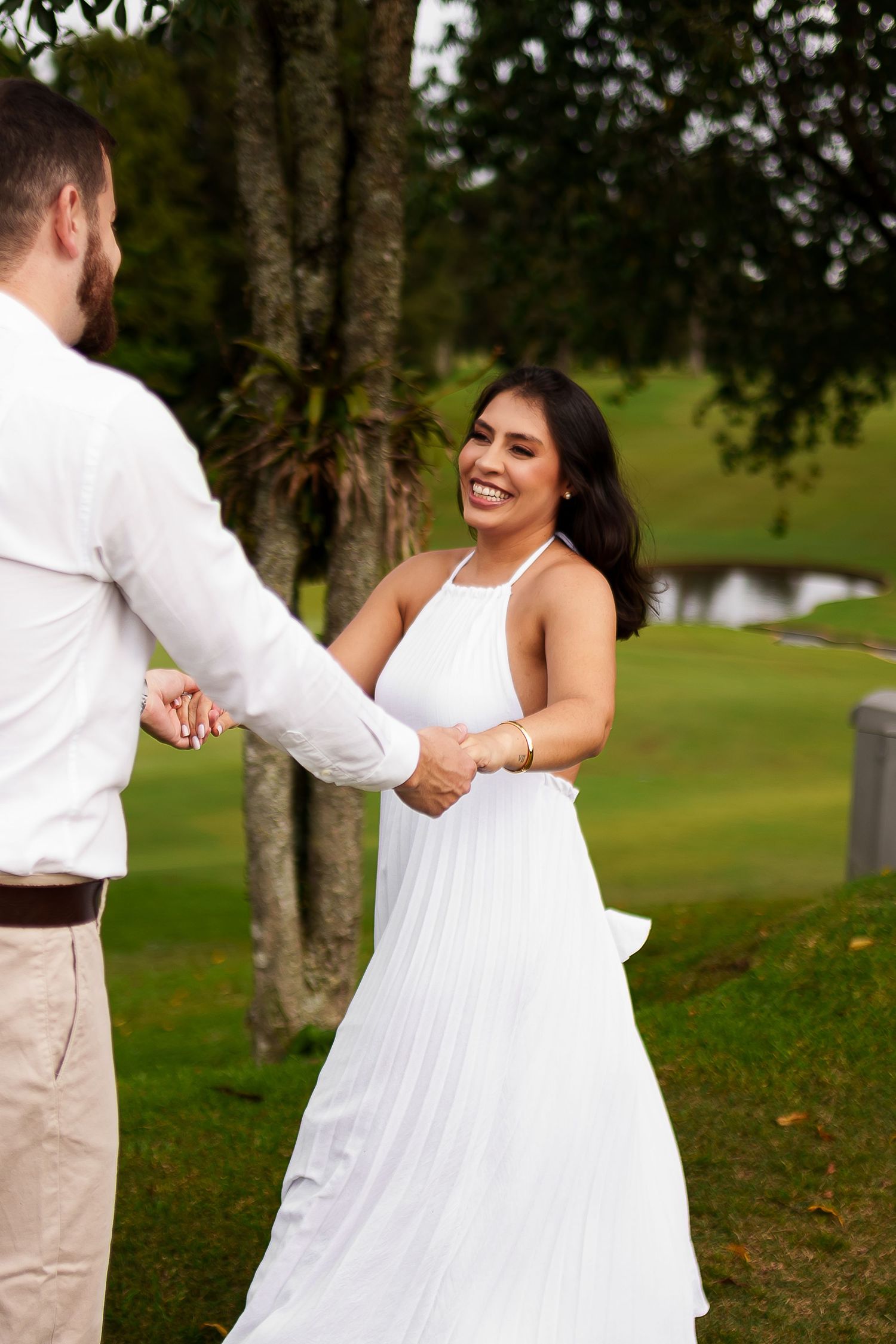 A smiling bride in a white halter dress holds hands with her groom outdoors near a tree and pond.