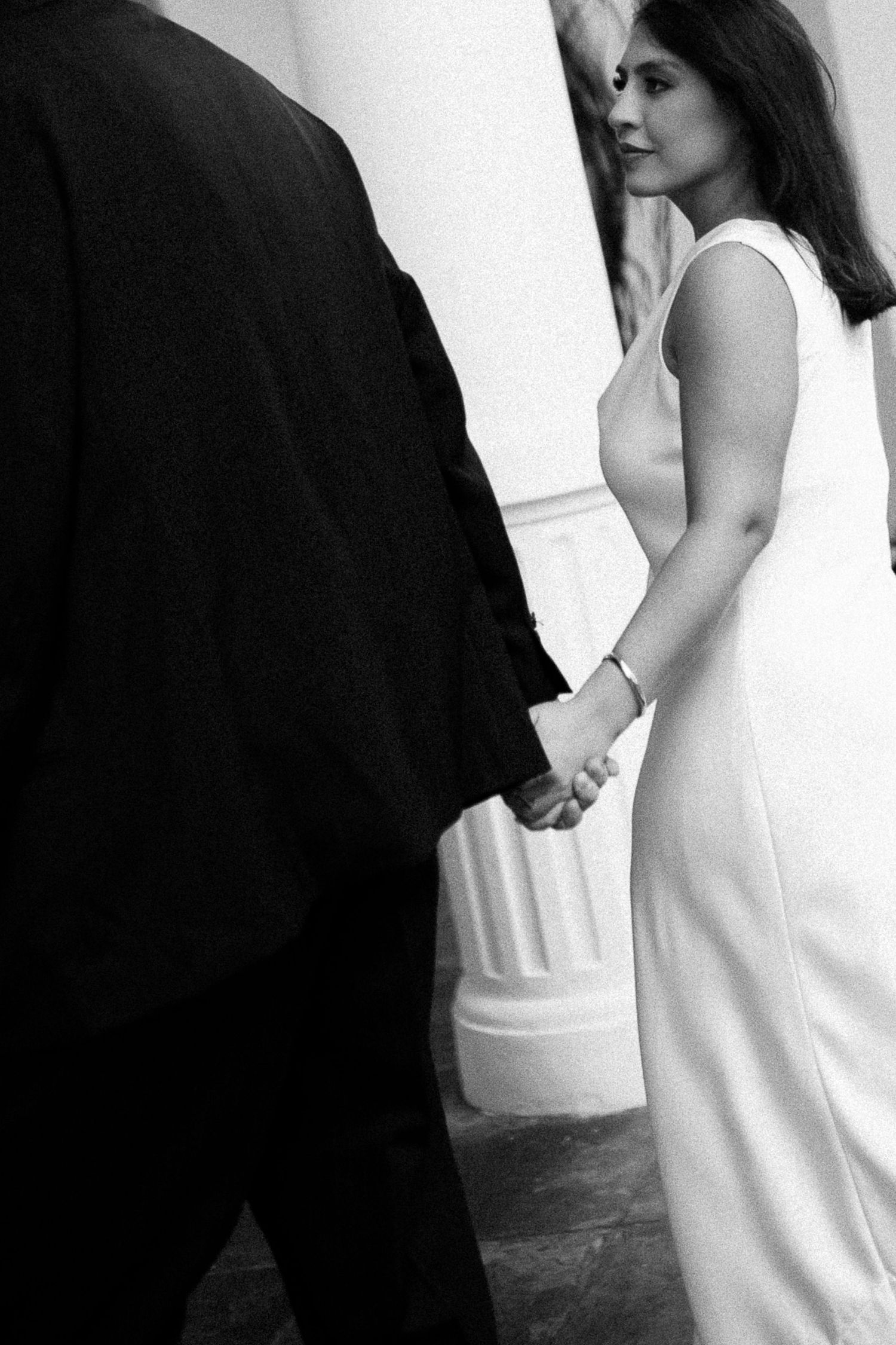 Black and white photo of a bride in white gown holding hands with a man in a dark suit near elegant columns.