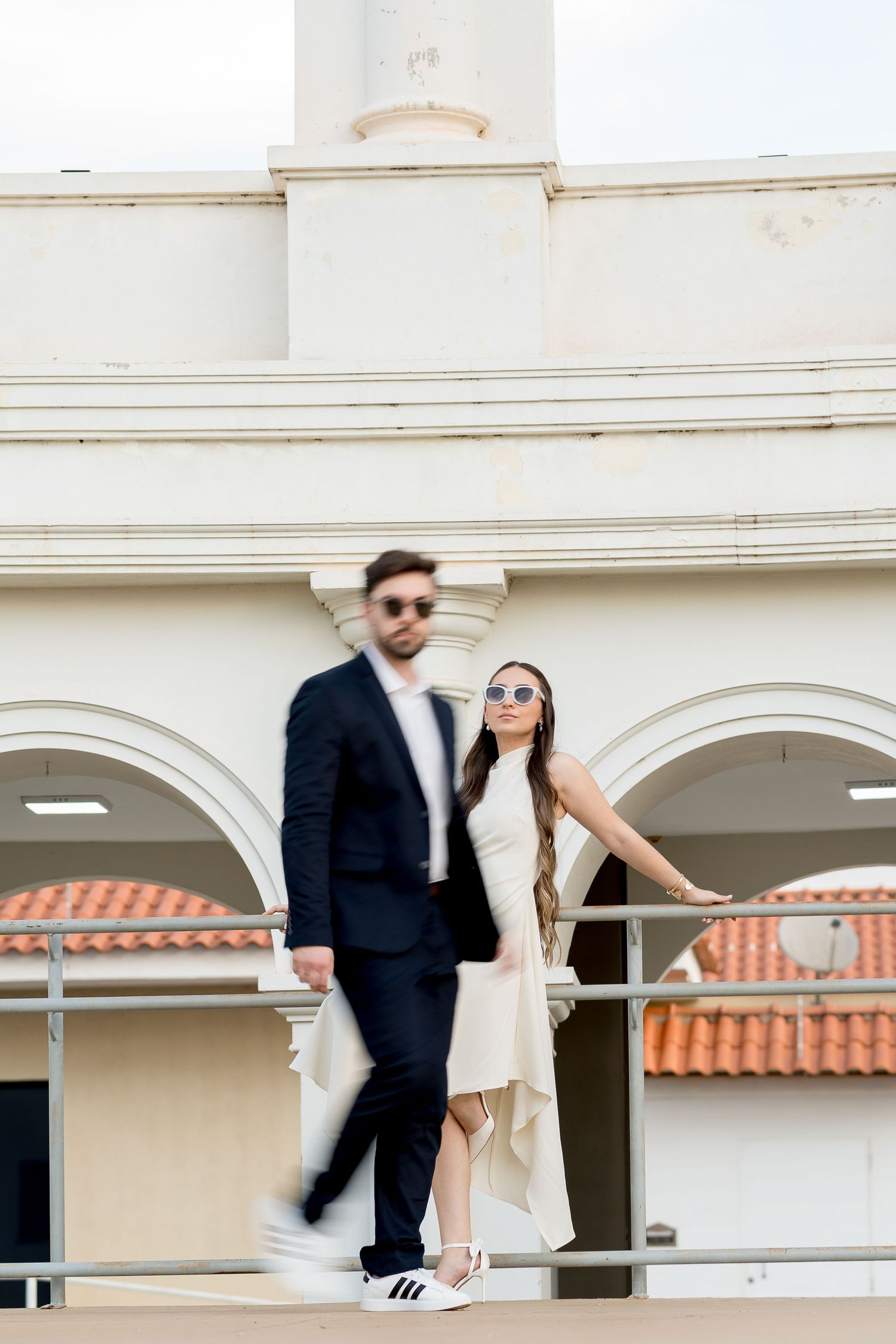 Stylish couple in sunglasses posing on a balcony, man in dark suit, woman in white dress with sneakers.