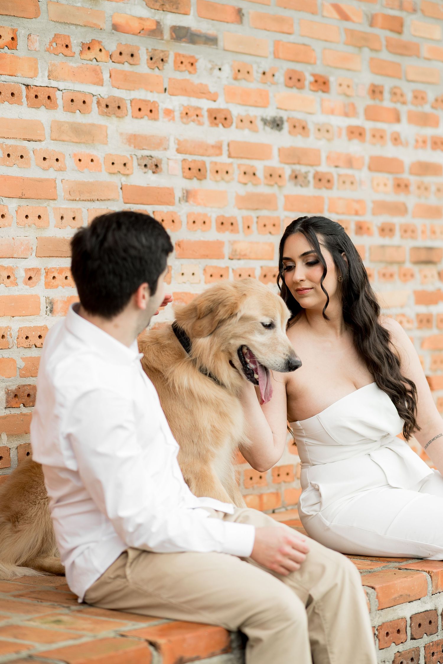 Bride in white dress and groom in white shirt petting a golden retriever against a rustic brick wall.