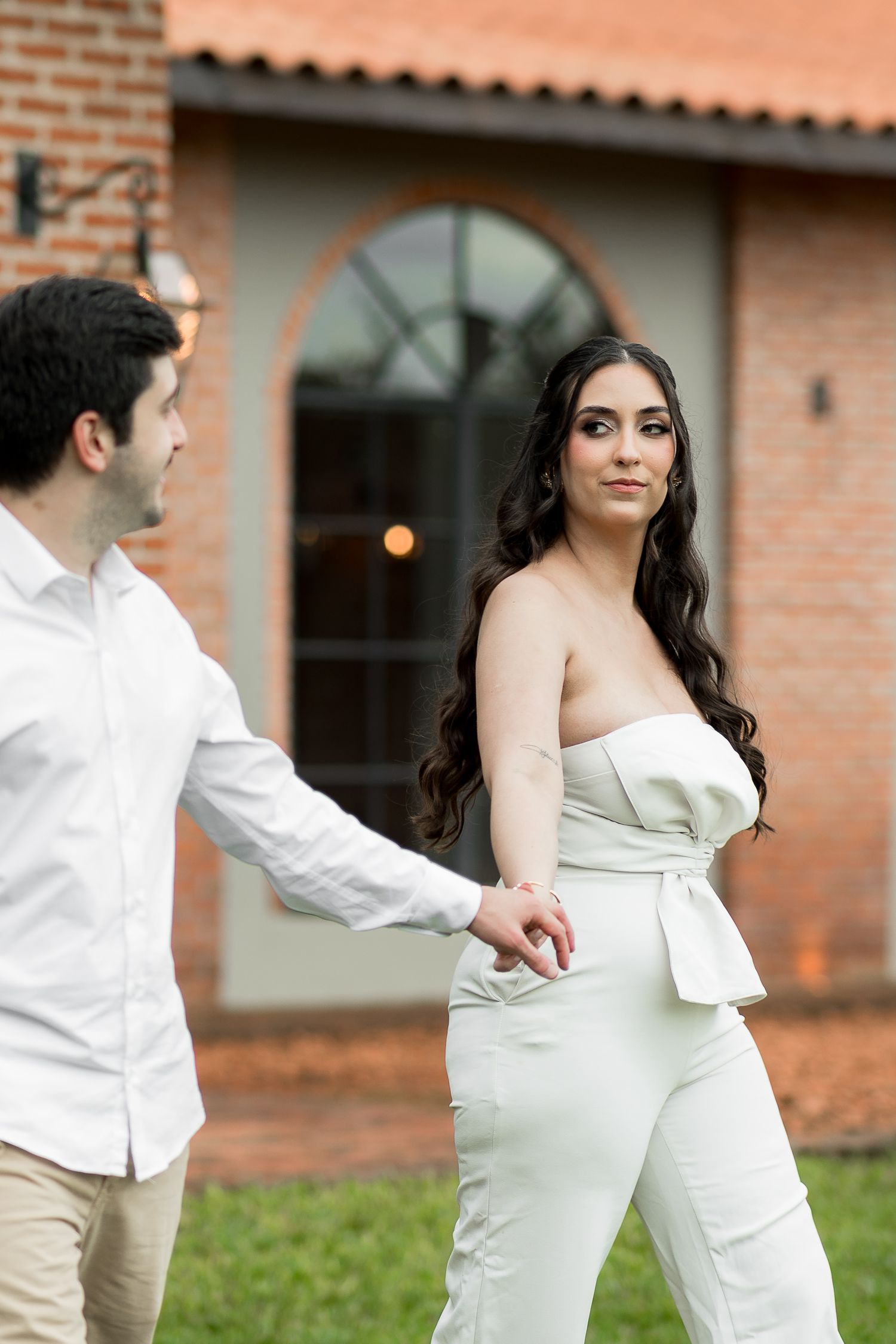 A couple holds hands outdoors, woman in white strapless jumpsuit with wavy hair, man in white shirt, brick building behind them.
