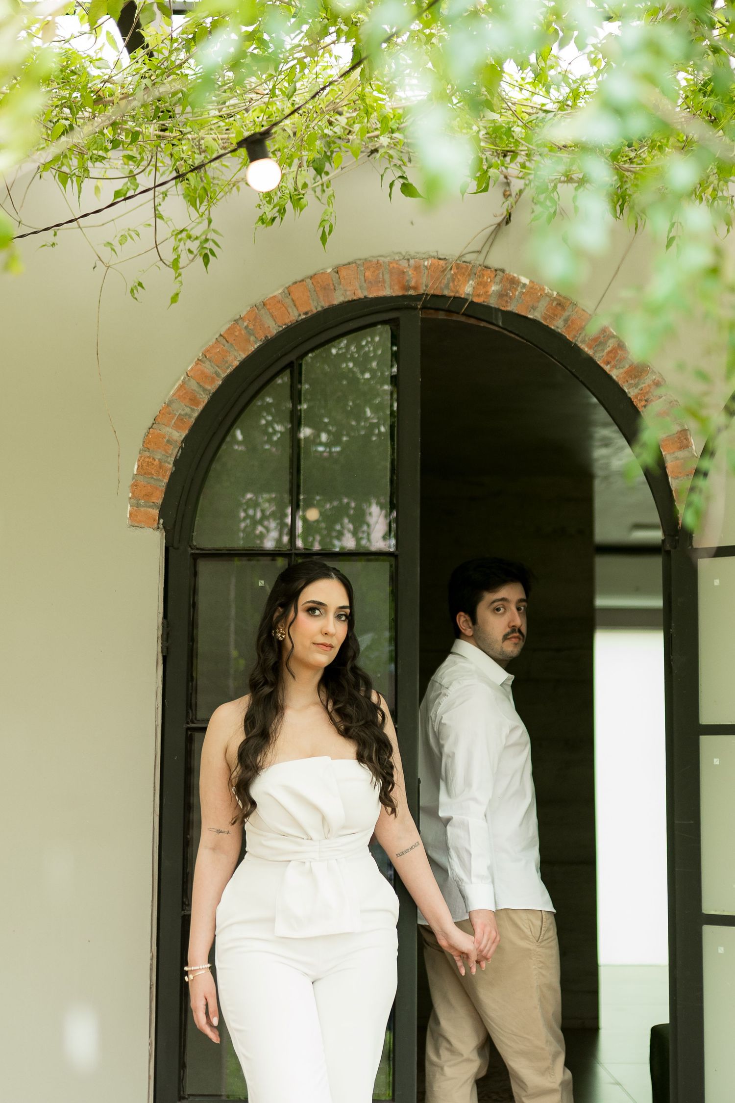Couple in white outfits posing under a brick arch doorway surrounded by lush green foliage.