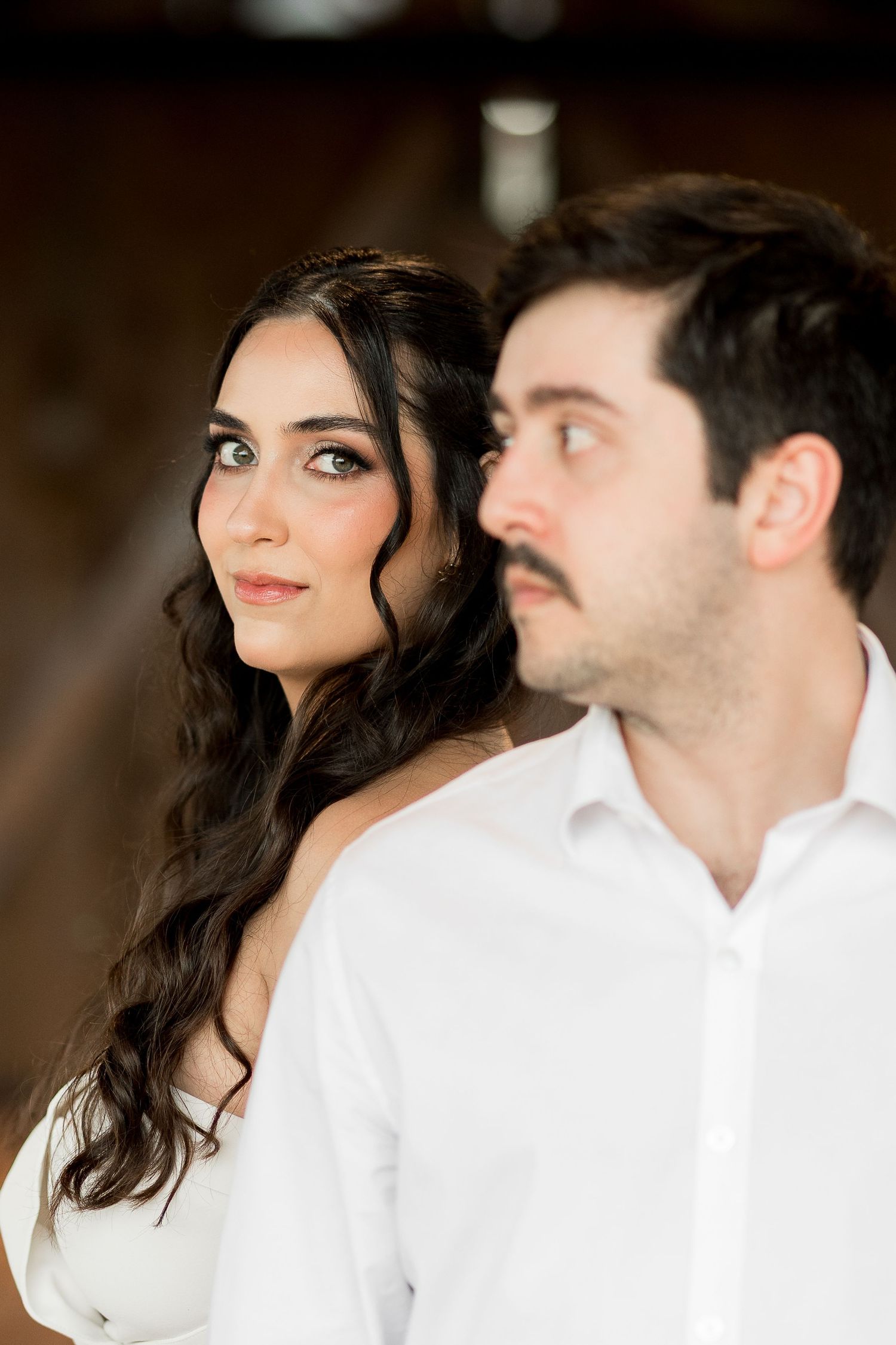 A bride with long dark wavy hair in a white dress gazes upward beside a man with a mustache in a white shirt.