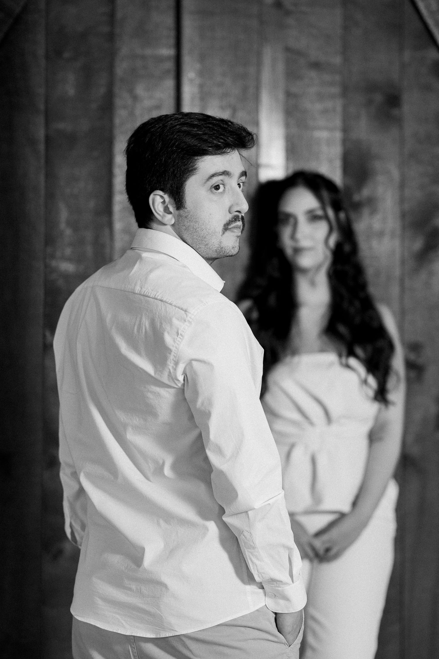 Black and white photo of a man in white shirt glancing back at a woman in white dress standing by wooden wall.