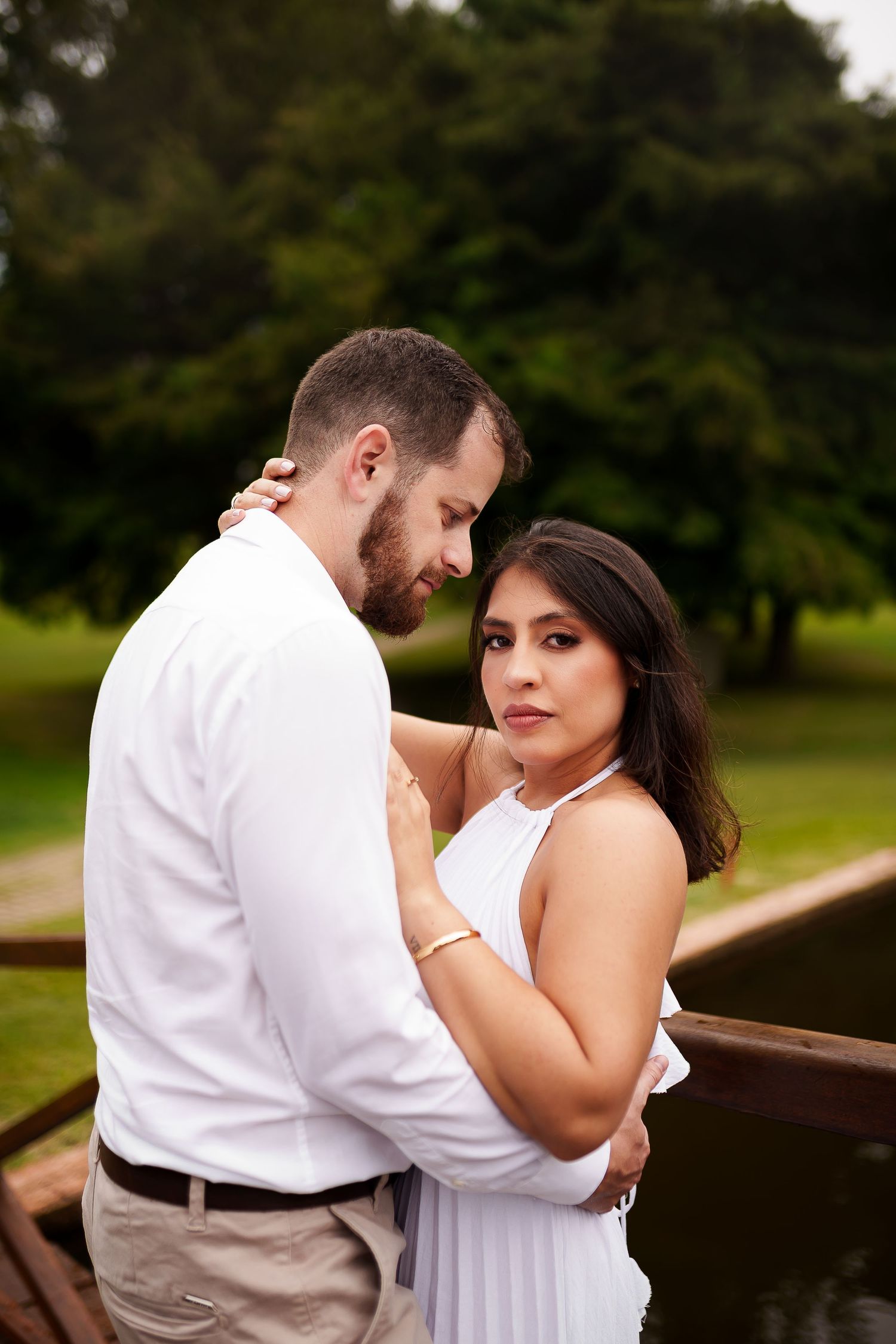 Couple in white outfits embracing romantically on a wooden bridge surrounded by lush green trees in a park.