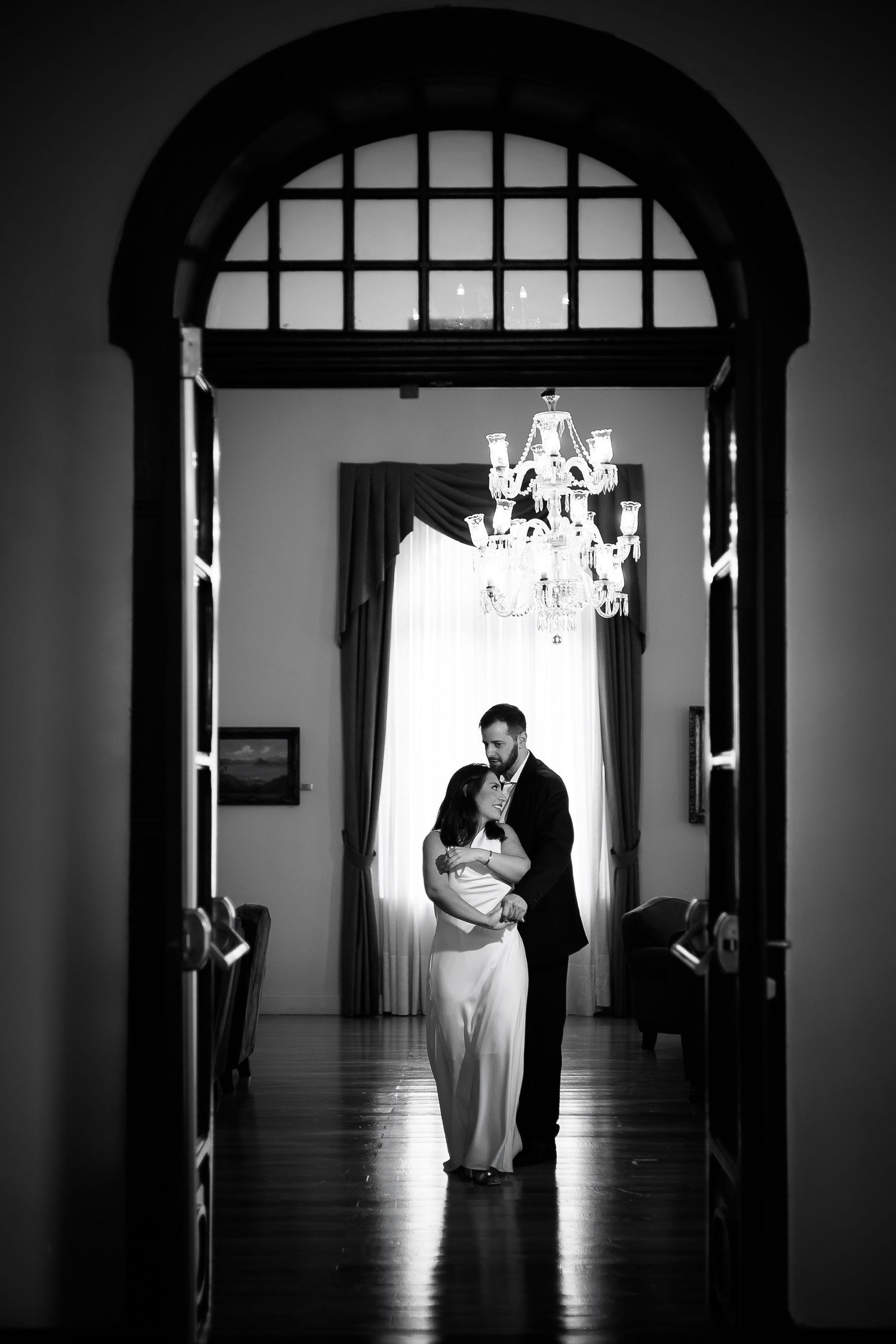 Black and white photo of a couple in formal attire embracing under a chandelier in an elegant arched doorway.