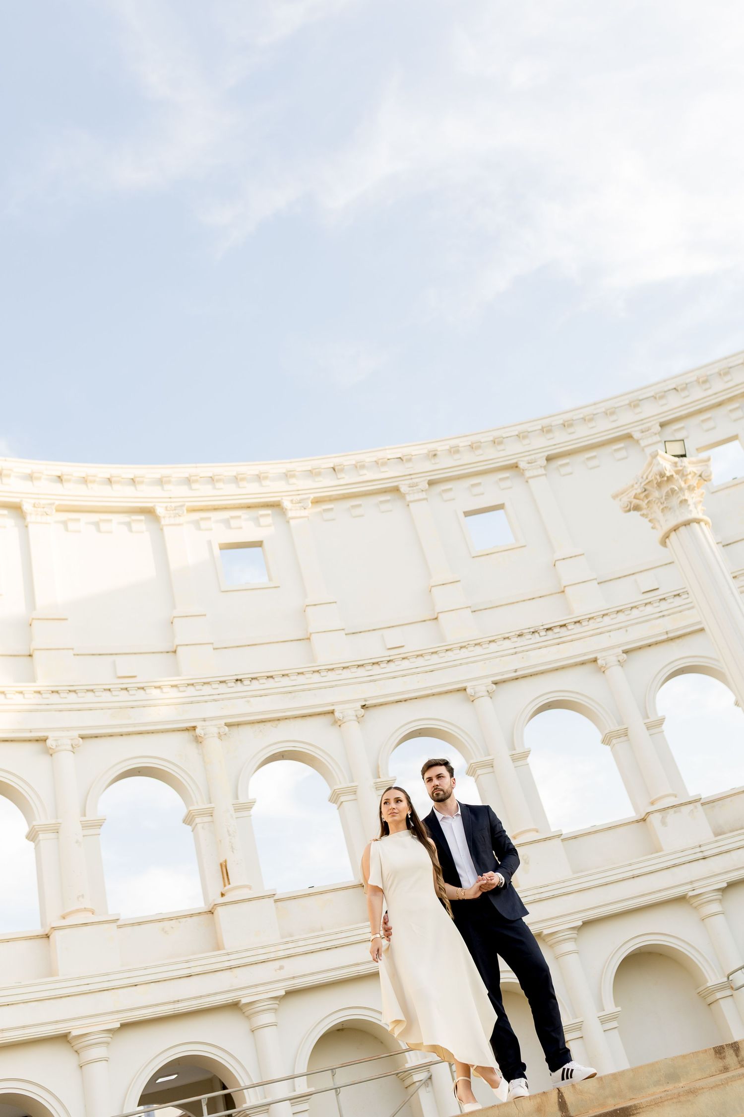 Elegant couple posing in front of the Colosseum in Rome, woman in white dress, man in navy suit.